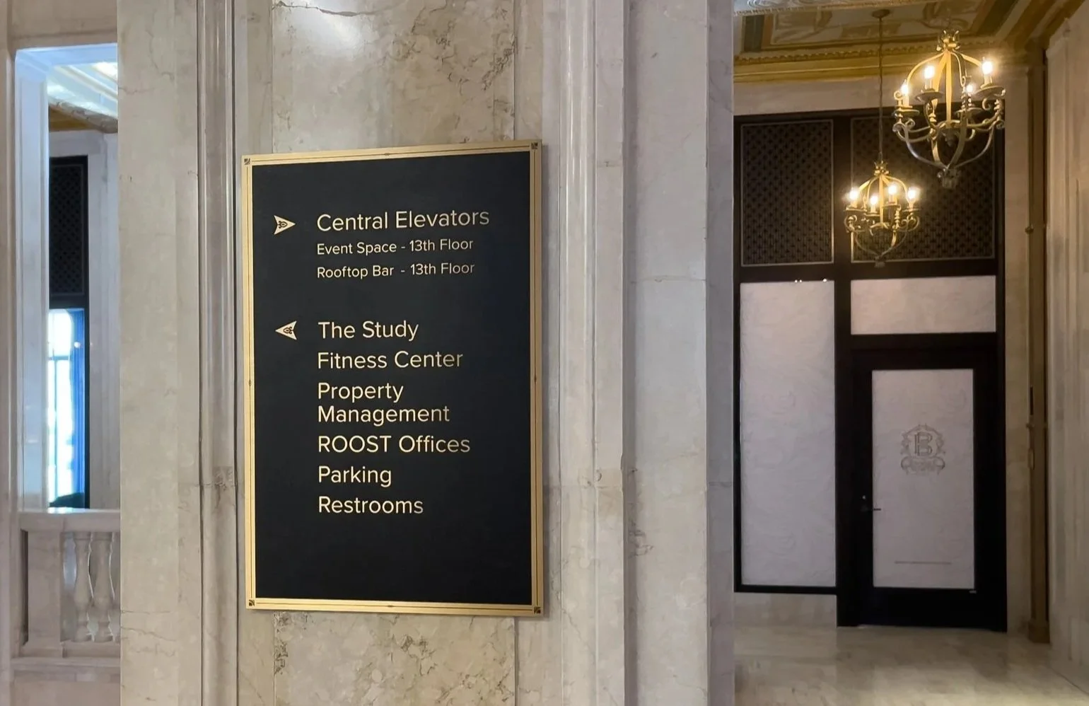 Sign listing directions to central elevators, event space, rooftop bar, the study, fitness center, property management, ROOST offices, parking, and restrooms inside a building with marble walls and ornate chandeliers.