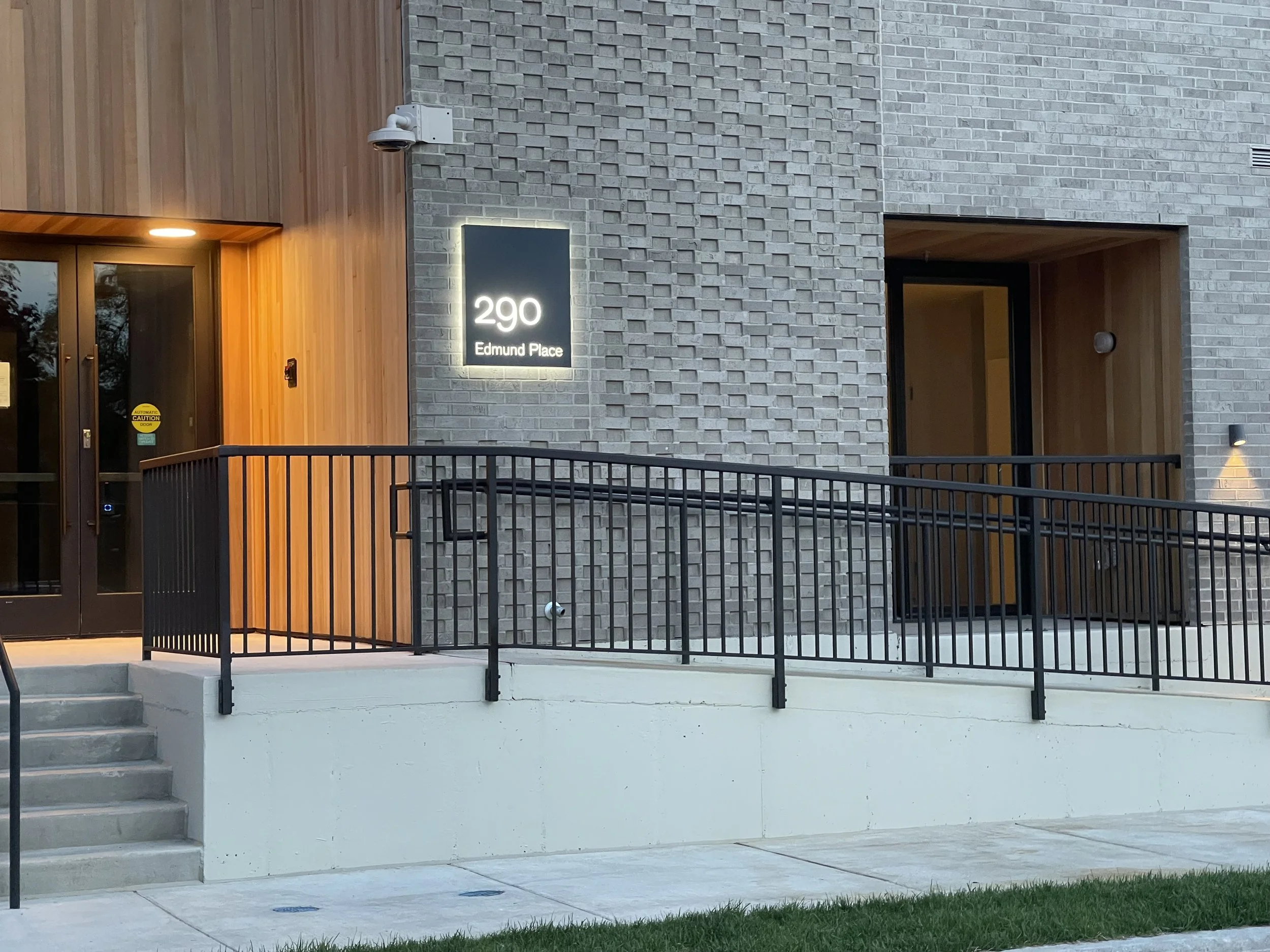 Exterior of a modern building with brick and wood siding, with street number 290 and address Edmund Place illuminated on a sign. The entrance has a staircase and an accessible ramp with black railings.