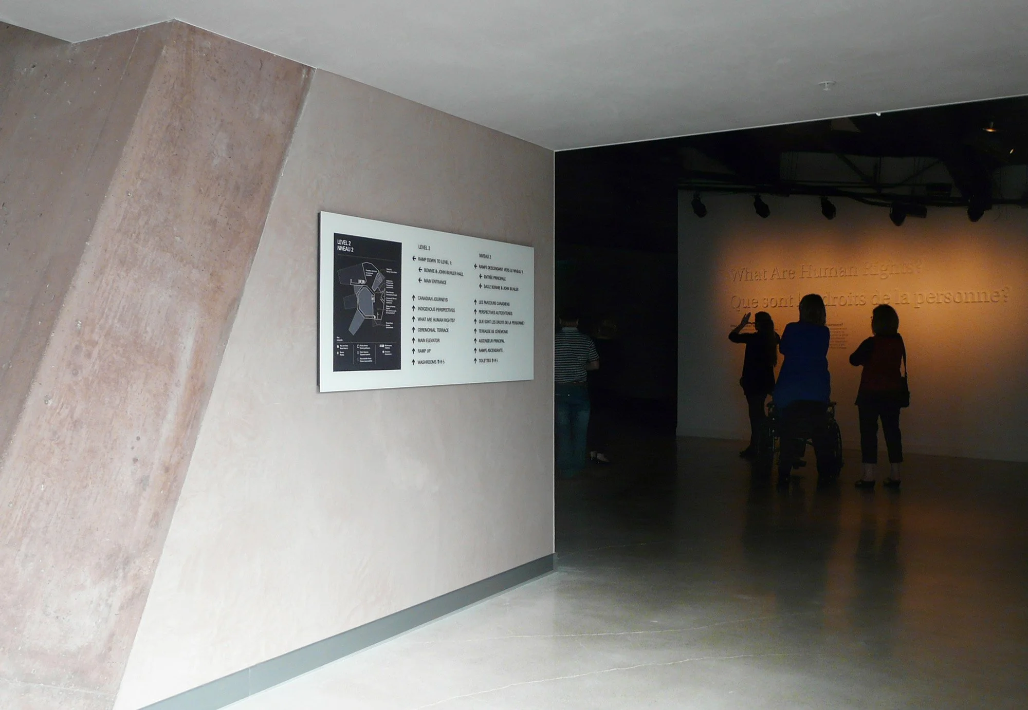 People silhouetted against a wall with a sign that reads 'What Are Human Rights?' in an indoor museum or exhibit space.