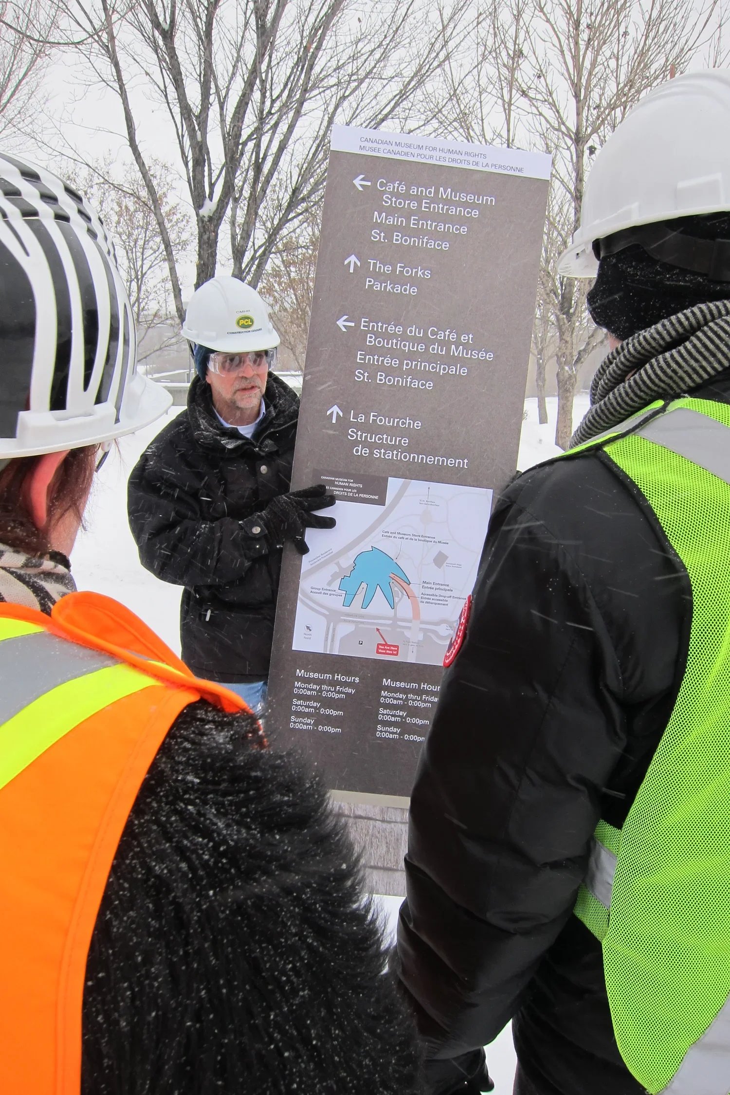 Group of construction workers wearing safety helmets and jackets, holding an information sign outdoors in a snowy environment.