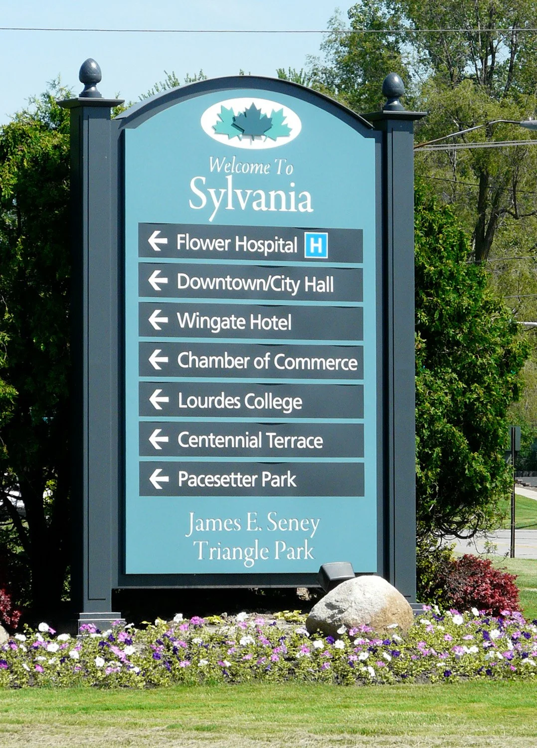 A welcome sign for Sylvania with directional signs pointing to various local locations such as Flower Hospital, Downtown/City Hall, Wingate Hotel, Chamber of Commerce, Lourdes College, Centennial Terrace, Pacesetter Park, and Triangle Park, surrounded by flowers and greenery.