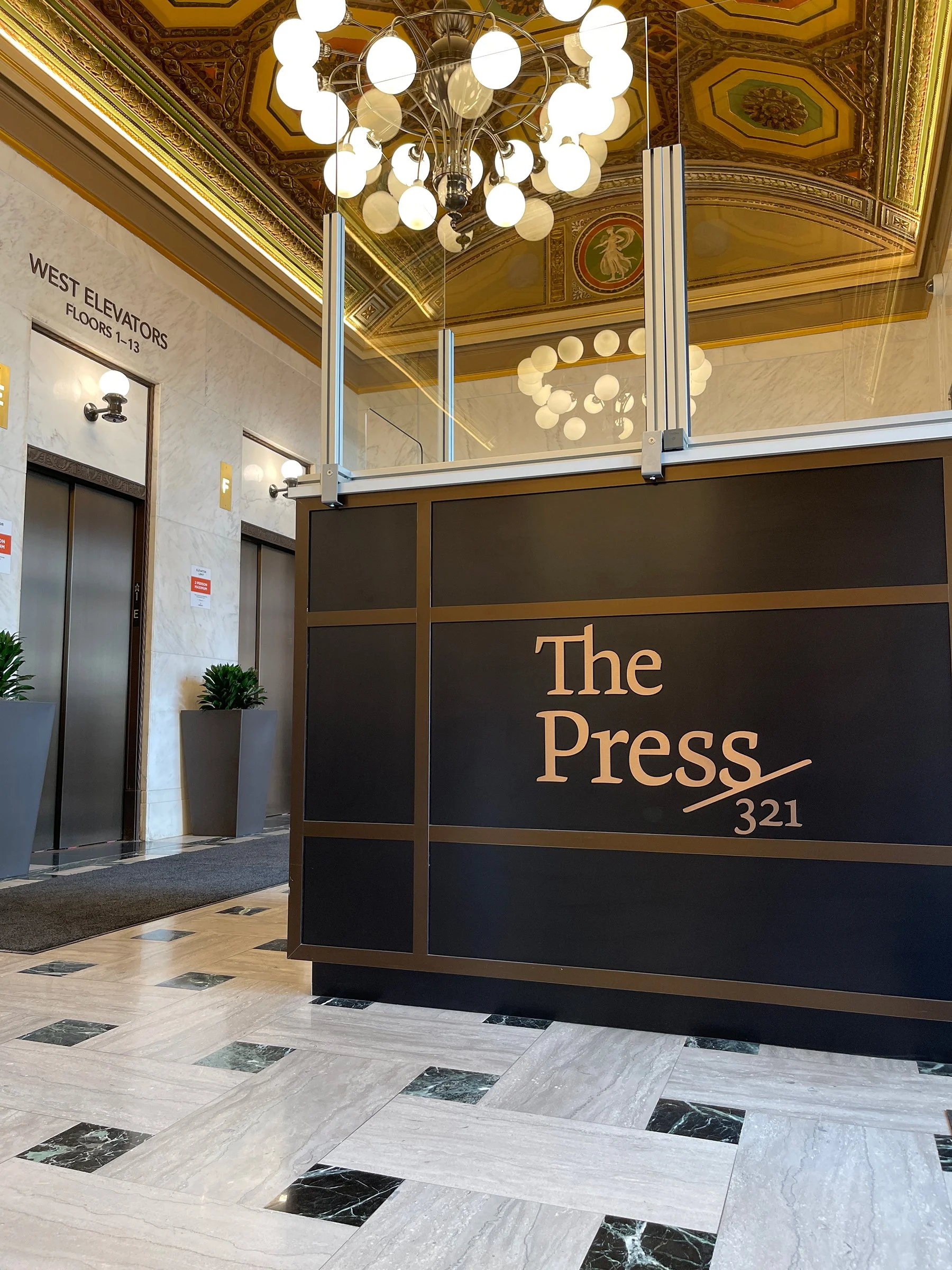 Lobby area with elevator doors on the left, potted plants, a decorative ornate ceiling with a chandelier, and a reception desk labeled 'The Press 321' in the foreground.
