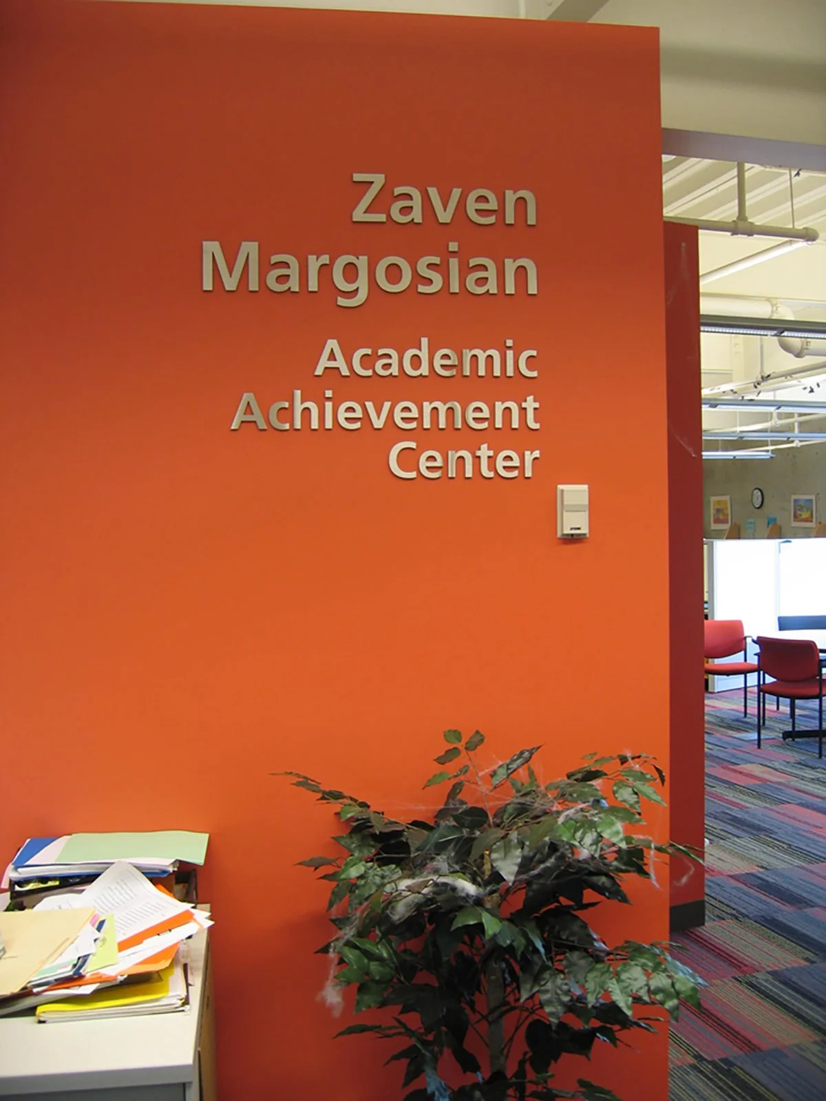 Orange wall with white letters reading 'Zaven Margosian Academic Achievement Center', a potted plant, and a table with scattered papers.