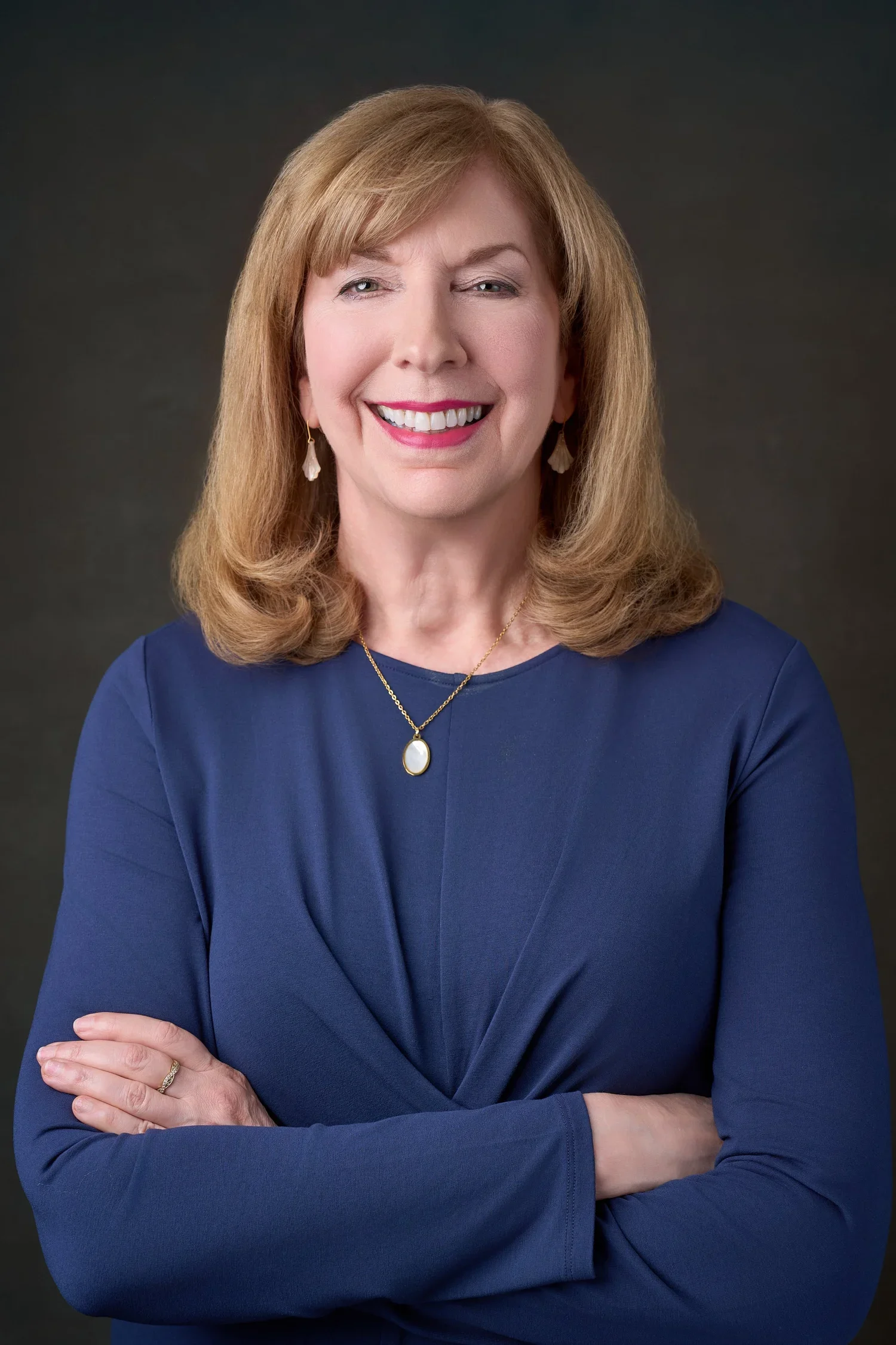 Portrait of a middle-aged woman with shoulder-length blonde hair, wearing a blue dress, gold jewelry, and smiling with arms crossed against a dark background.