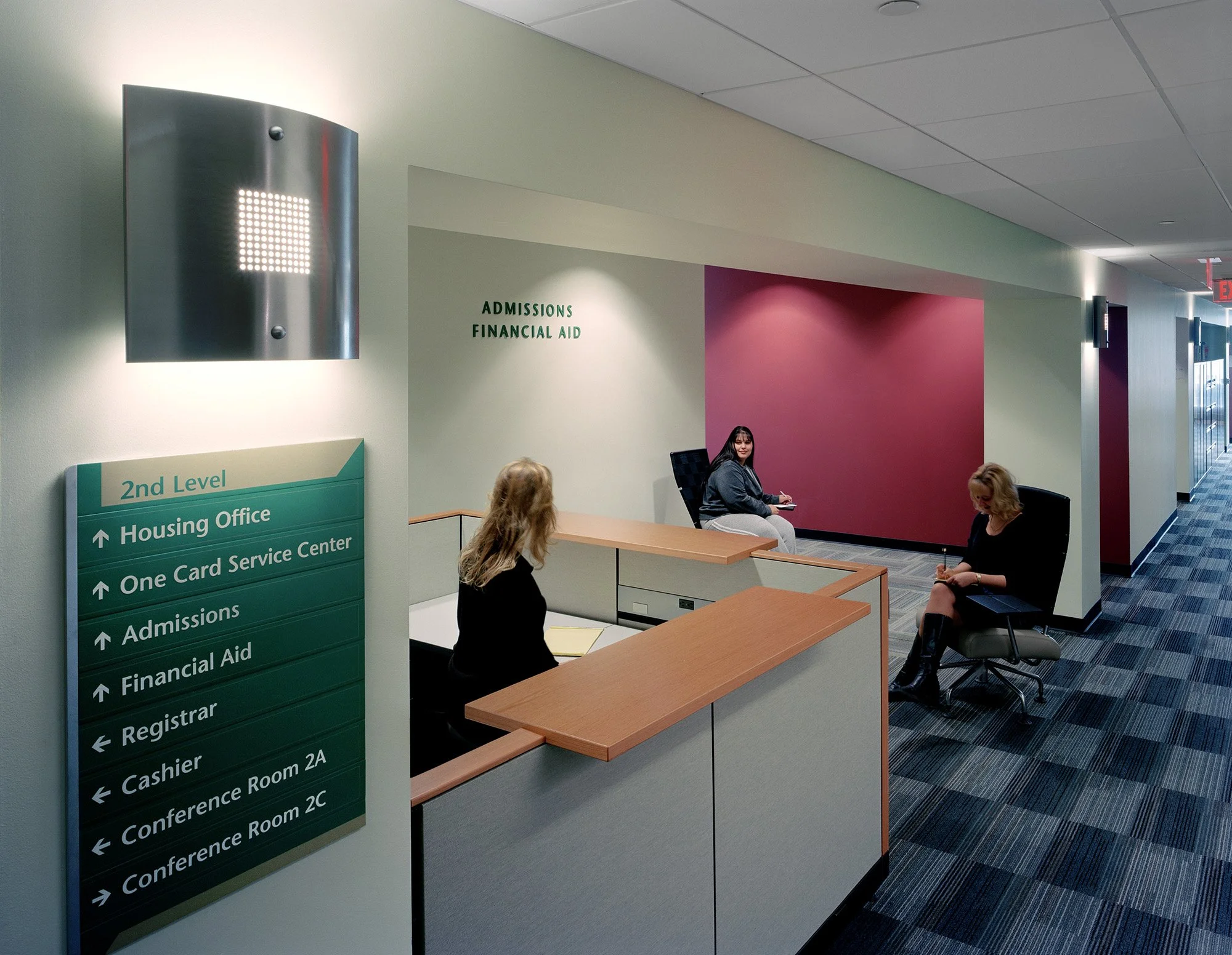 School or university reception area with three women sitting, sign indicating second level offices including housing, financial aid, admissions, registrar, cashier, and conference rooms, and a decorative wall with pink paint.
