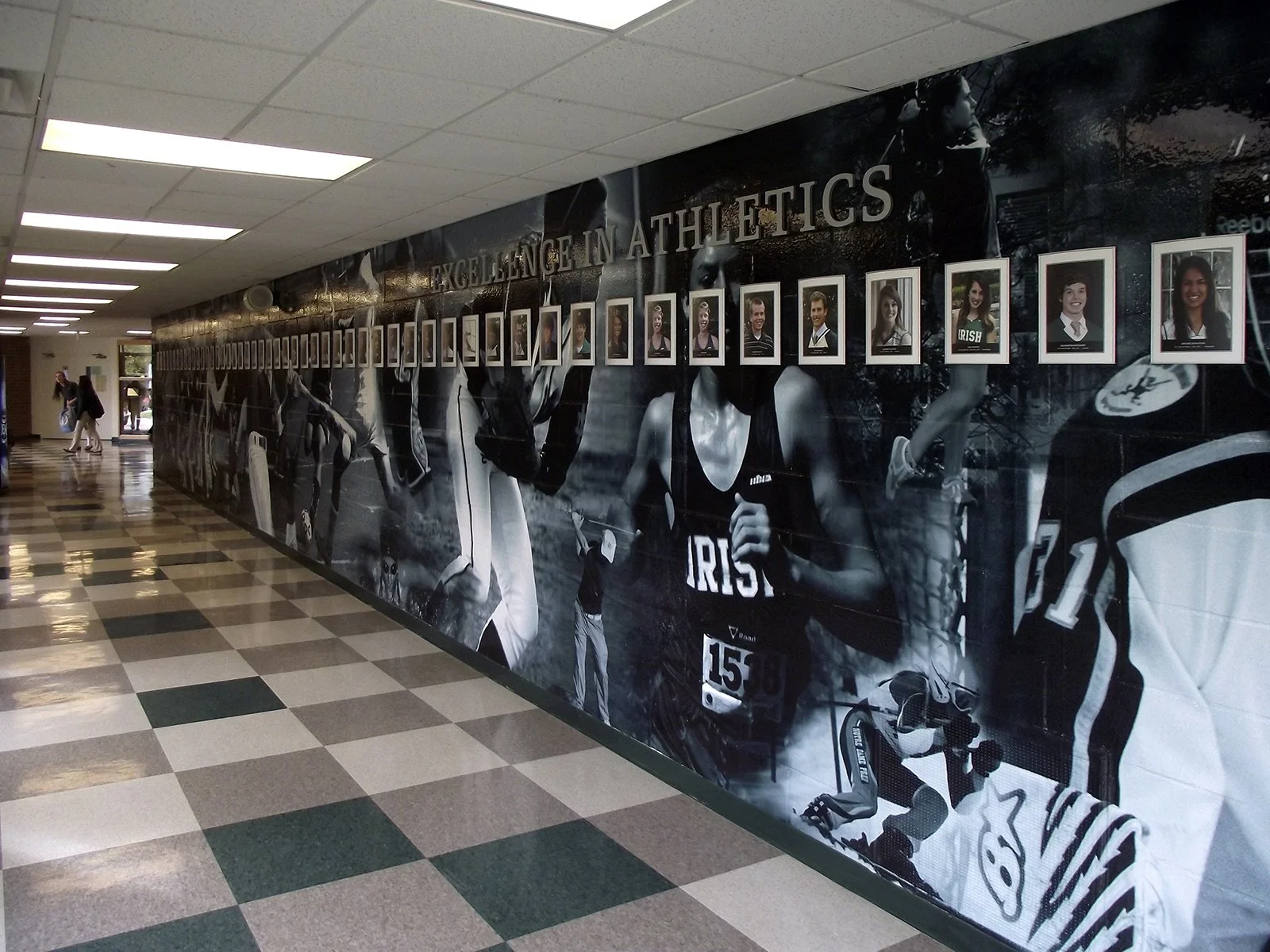 Hallway with a large black-and-white mural of athletes and framed photos of athletes on the wall, with the text "Excellence in Athletics."
