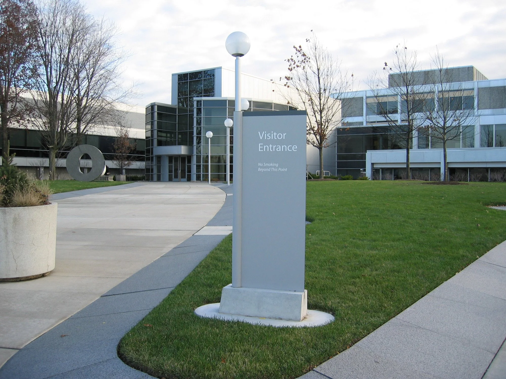 Modern office building with a sign that says 'Visitor Entrance' and a note 'No Smoking Beyond This Point', surrounded by grass, trees, and a concrete walkway.