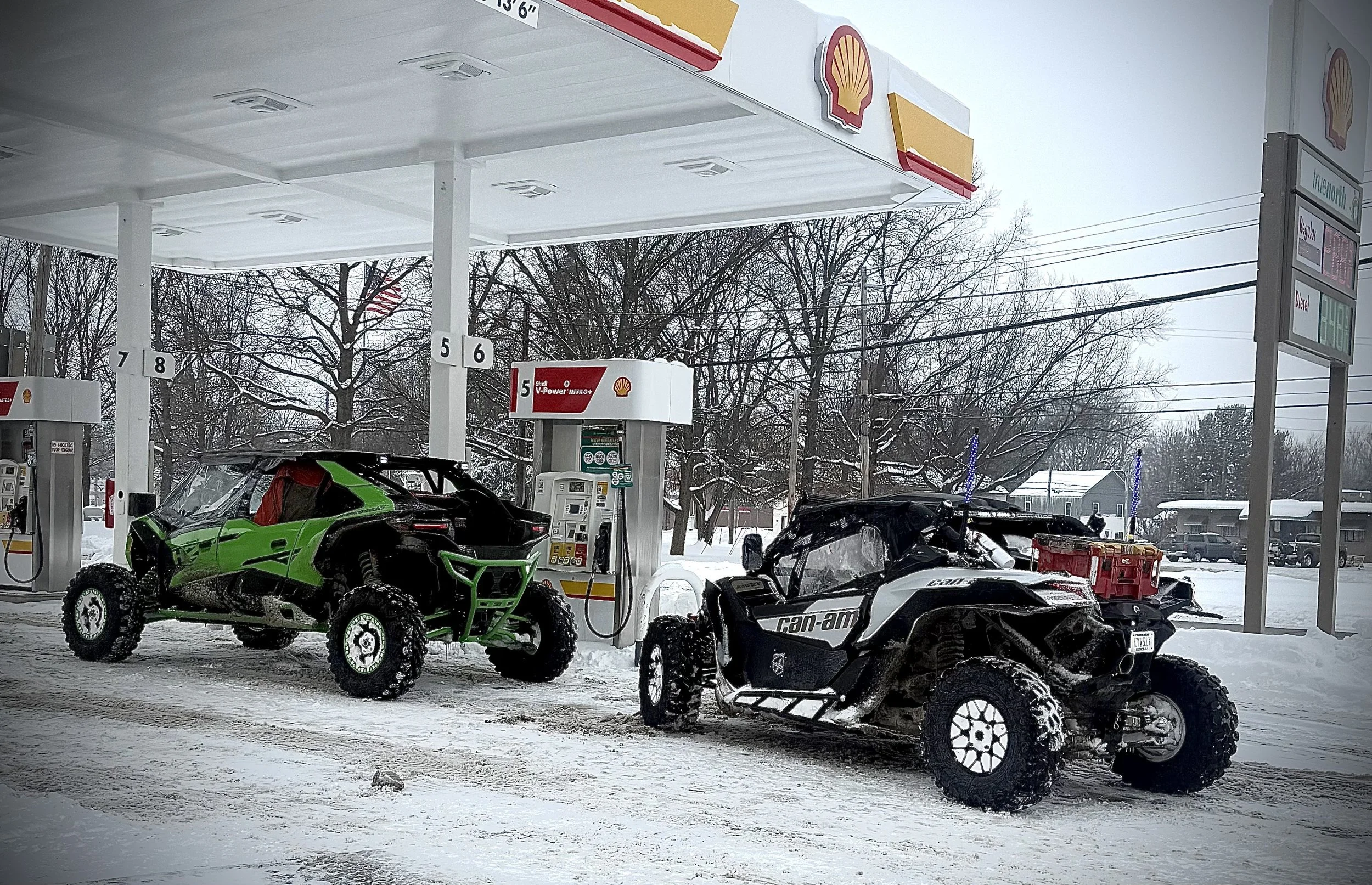 Two off-road vehicles, a green and black side-by-side and a white and black Can-Am off-road vehicle, parked at a snow-covered Shell gas station during winter.