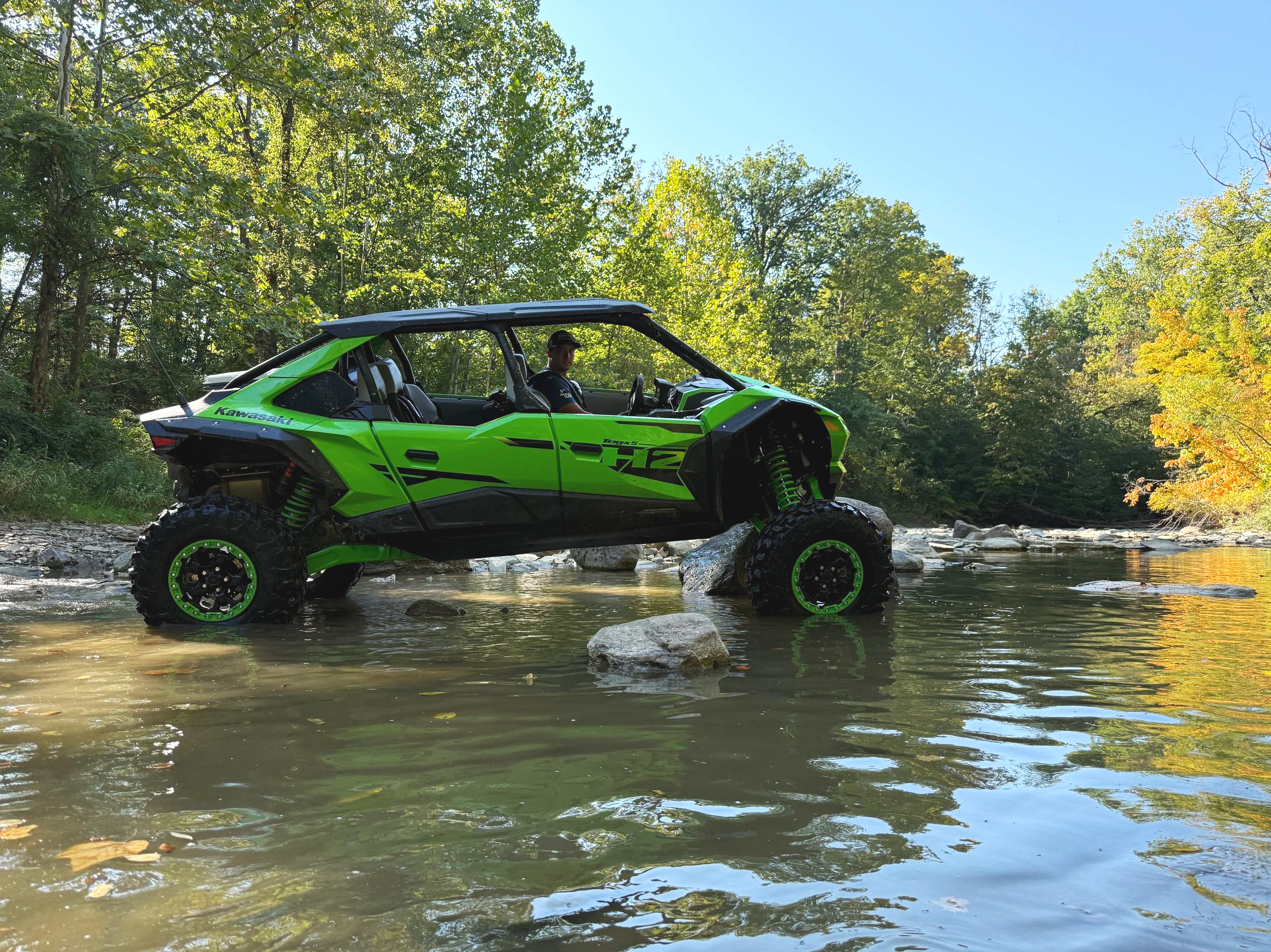 A green off-road vehicle crossing a shallow river in a wooded area with trees on either side and clear blue sky.