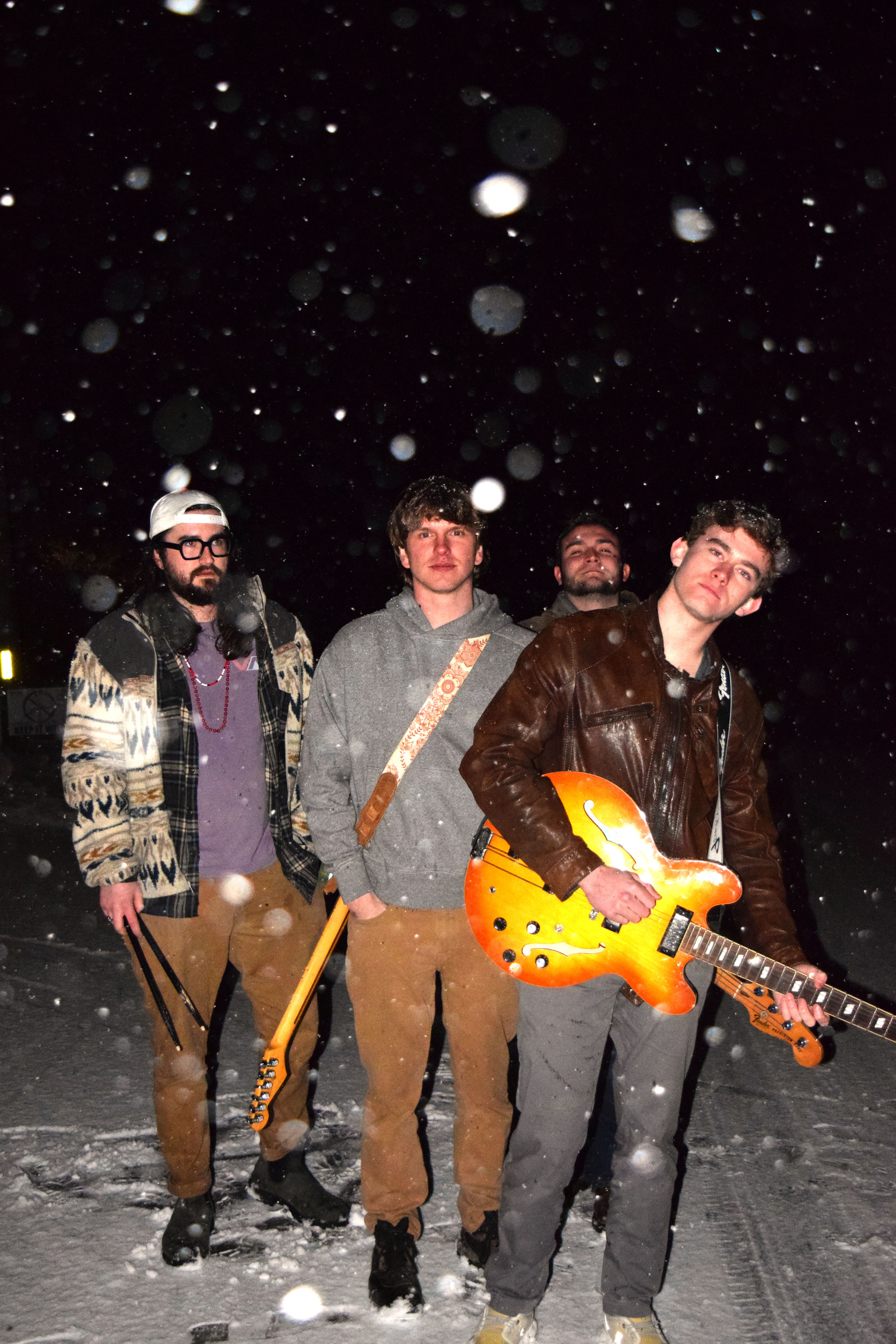 Four young men standing outdoors at night in falling snow, with one holding a colorful electric guitar.
