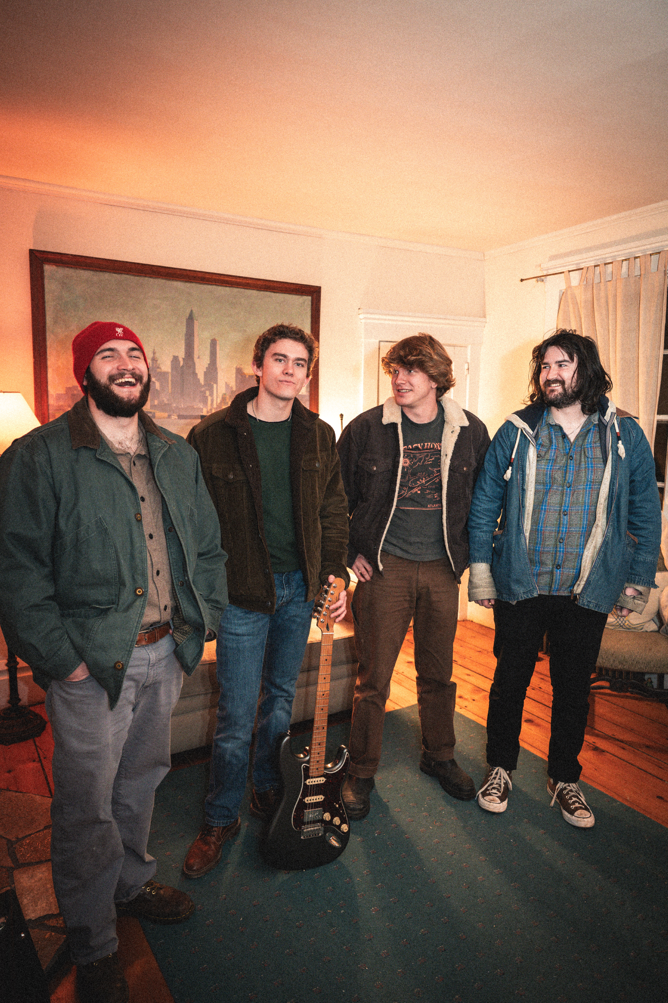 Four young men standing together indoors, with a guitar on the floor in front of them, smiling and looking at the camera. The room has wooden floors, a painting of a city skyline on the wall, and curtains on the window.