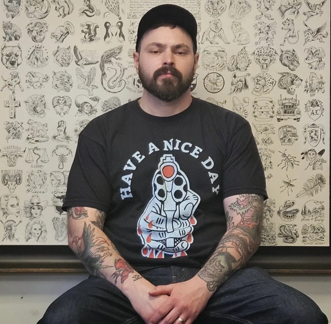 A man with a beard wearing a black baseball cap and a black T-shirt with a graphic of a tattooed hand holding a gun and the text "Have a Nice Day". He has tattoos on his arms and is sitting with his hands clasped in front of him. Behind him is a wall decorated with various tattoo-style drawings and designs.