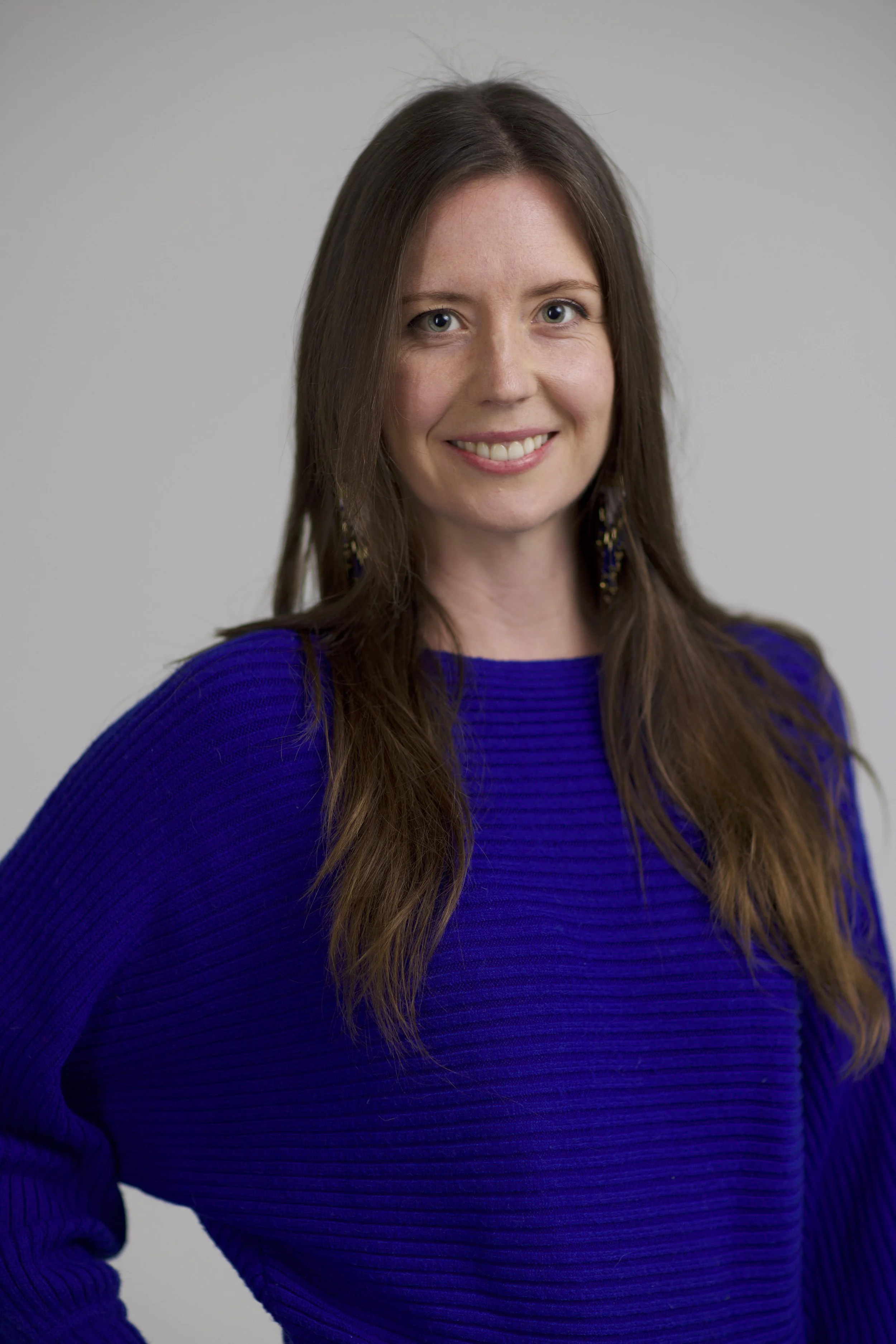 A woman with long brown hair, blue eyes, and a warm smile, wearing a royal blue sweater and dangling earrings, standing against a plain light gray background.