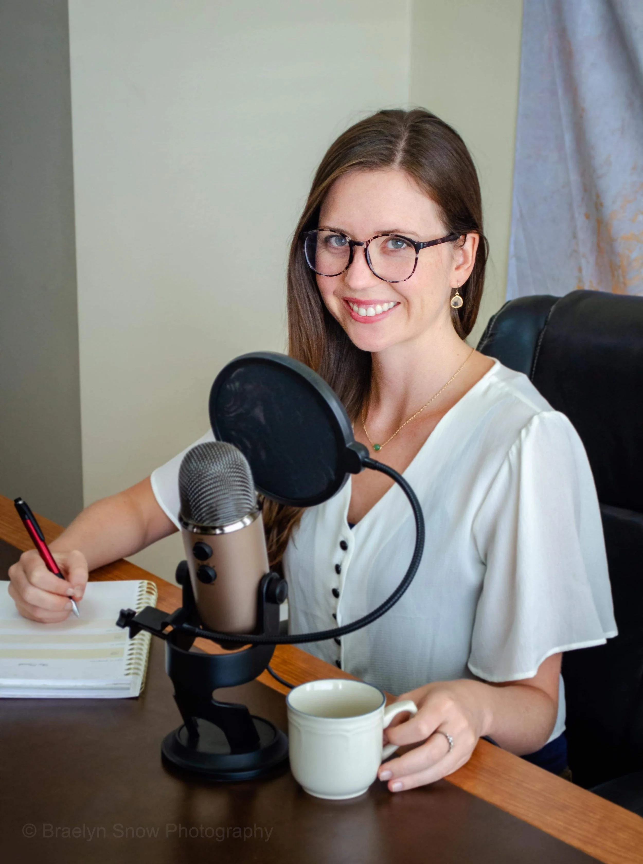 A woman with glasses and long brown hair smiling at a microphone, sitting at a desk with a mug, notebook, and pen.