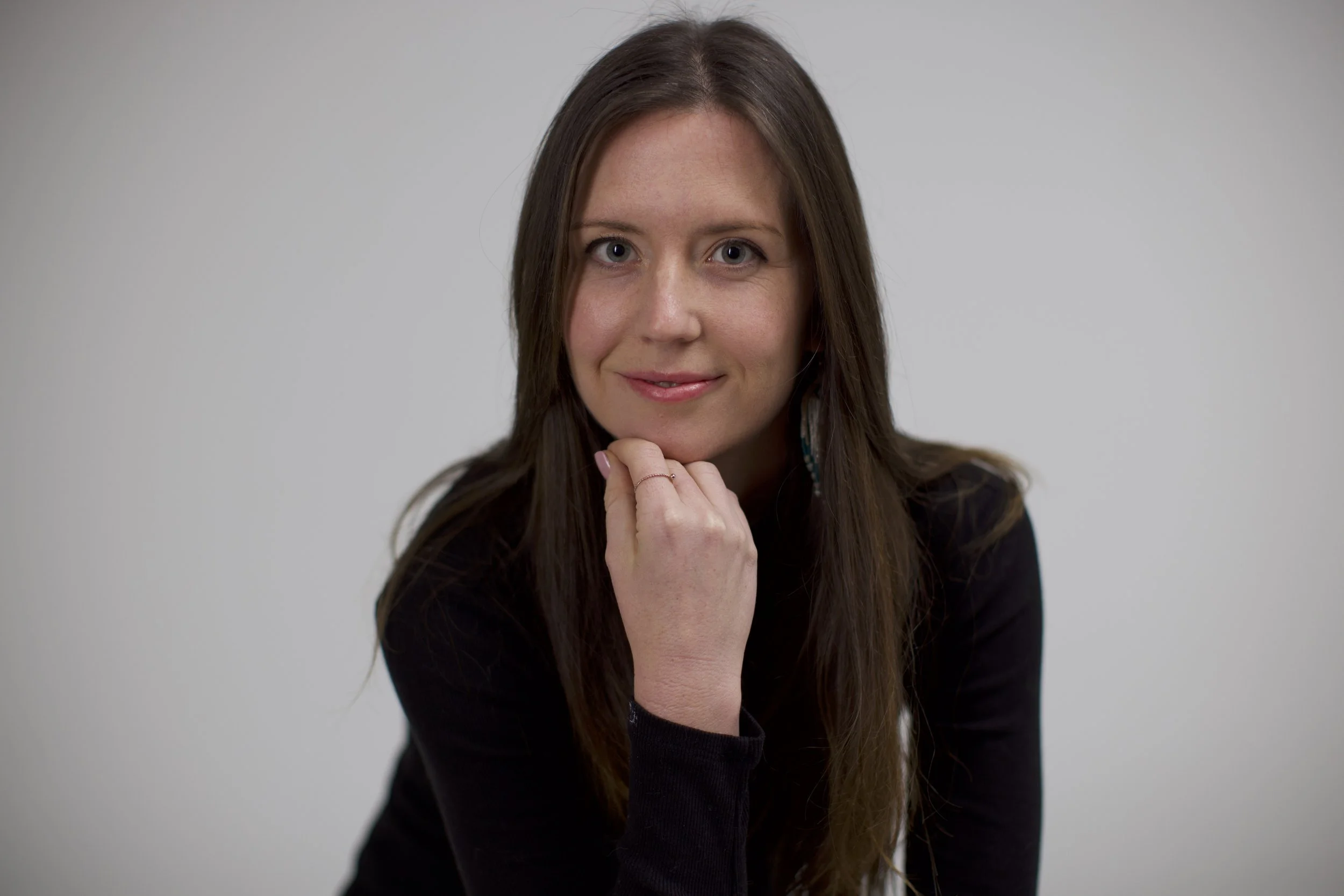 A woman with long brown hair smiling at the camera, resting her chin on her left hand against a plain light gray background.