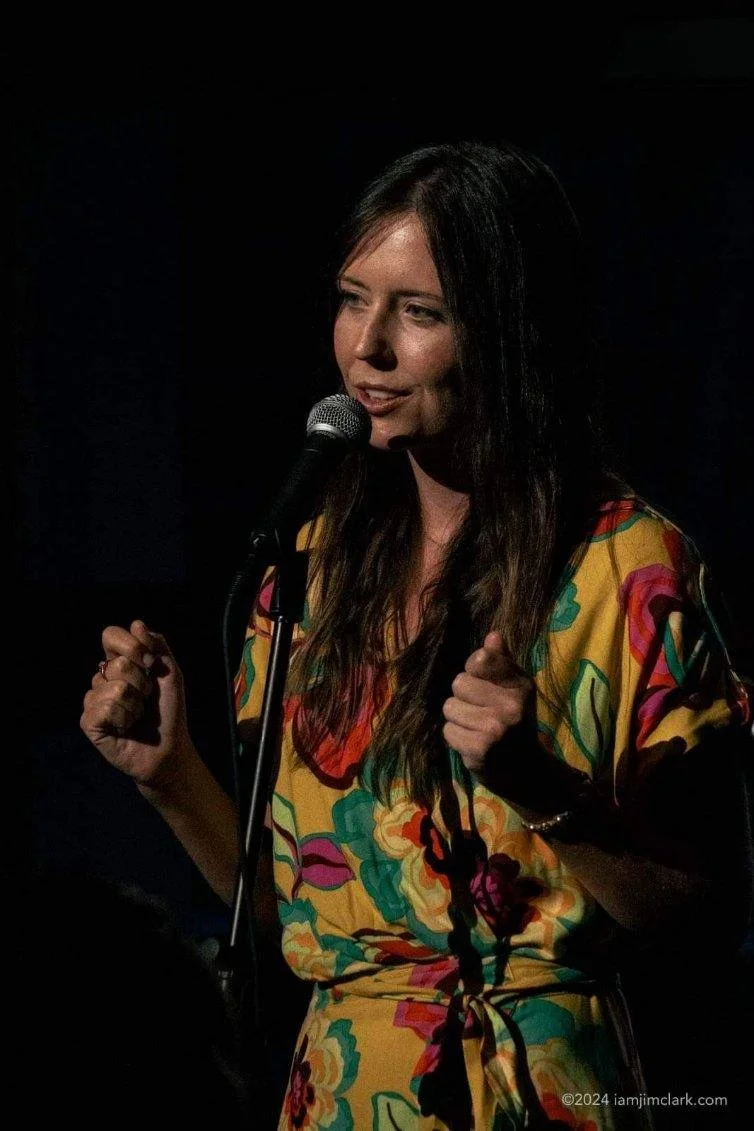 A woman with long dark hair performing stand-up comedy on stage, holding a microphone, wearing a colorful, floral-patterned dress.