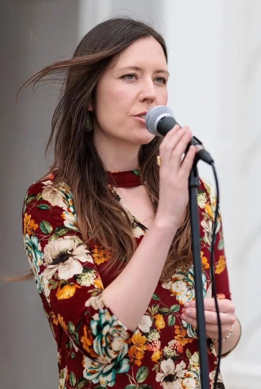 A woman with long brown hair singing into a microphone, wearing a floral dress with a choker.