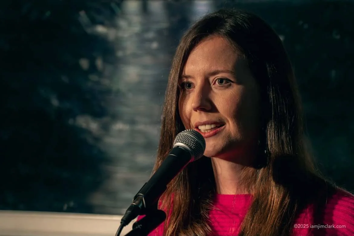 A woman with long brown hair speaking into a microphone on stage, wearing a pink top.