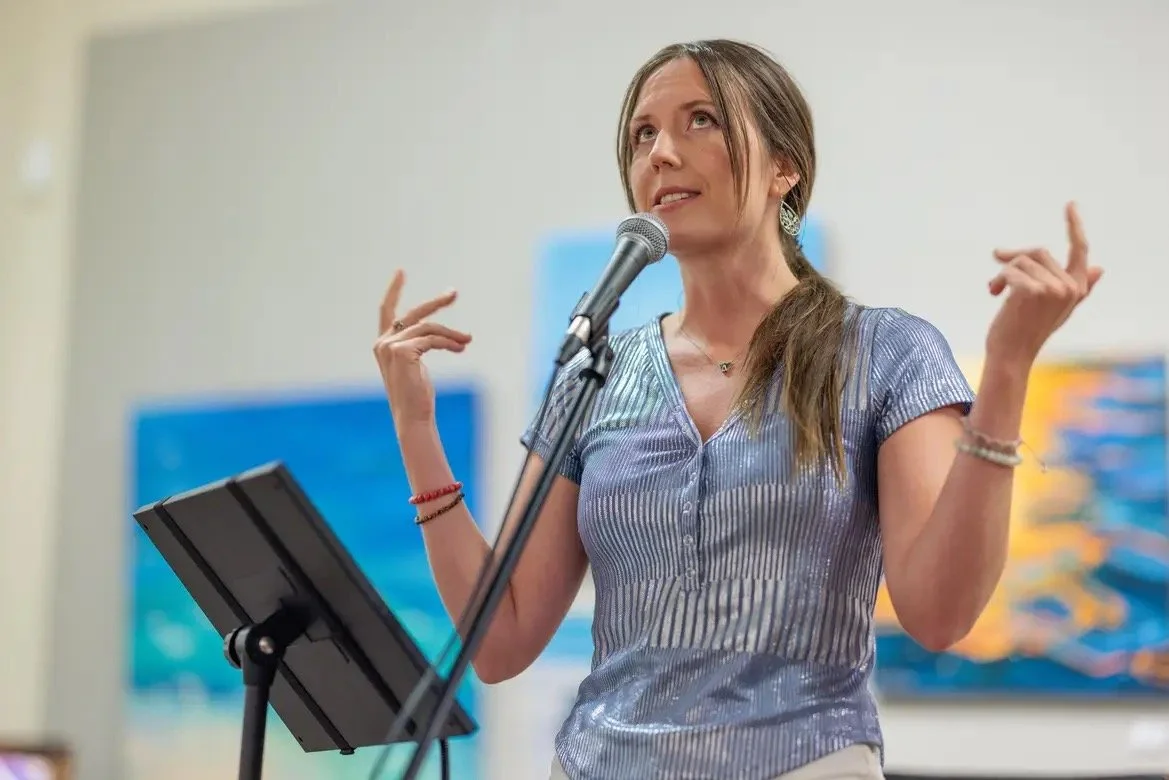 A woman standing at a podium with a microphone, gesturing with her hands while speaking.