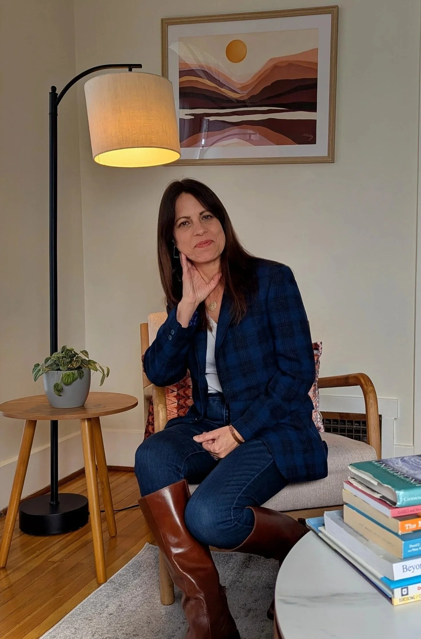A woman sitting on a chair in a cozy living room, wearing a plaid blazer, jeans, and brown boots, with her hand resting on her face.