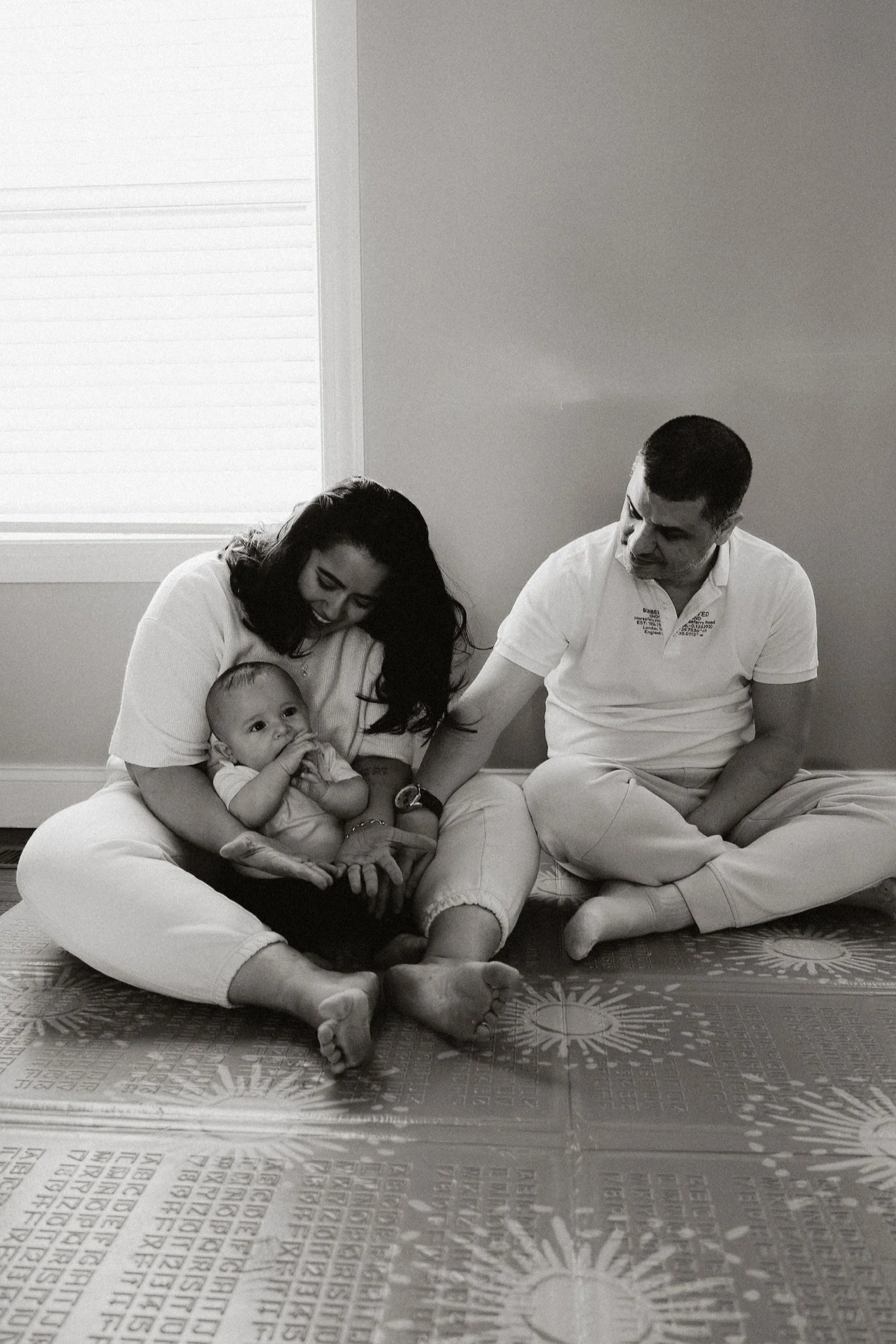Black and white photo of a family sitting on the floor indoors. A woman holds a baby, and a man is sitting beside them. The woman is smiling down at the baby, who is looking at the camera.