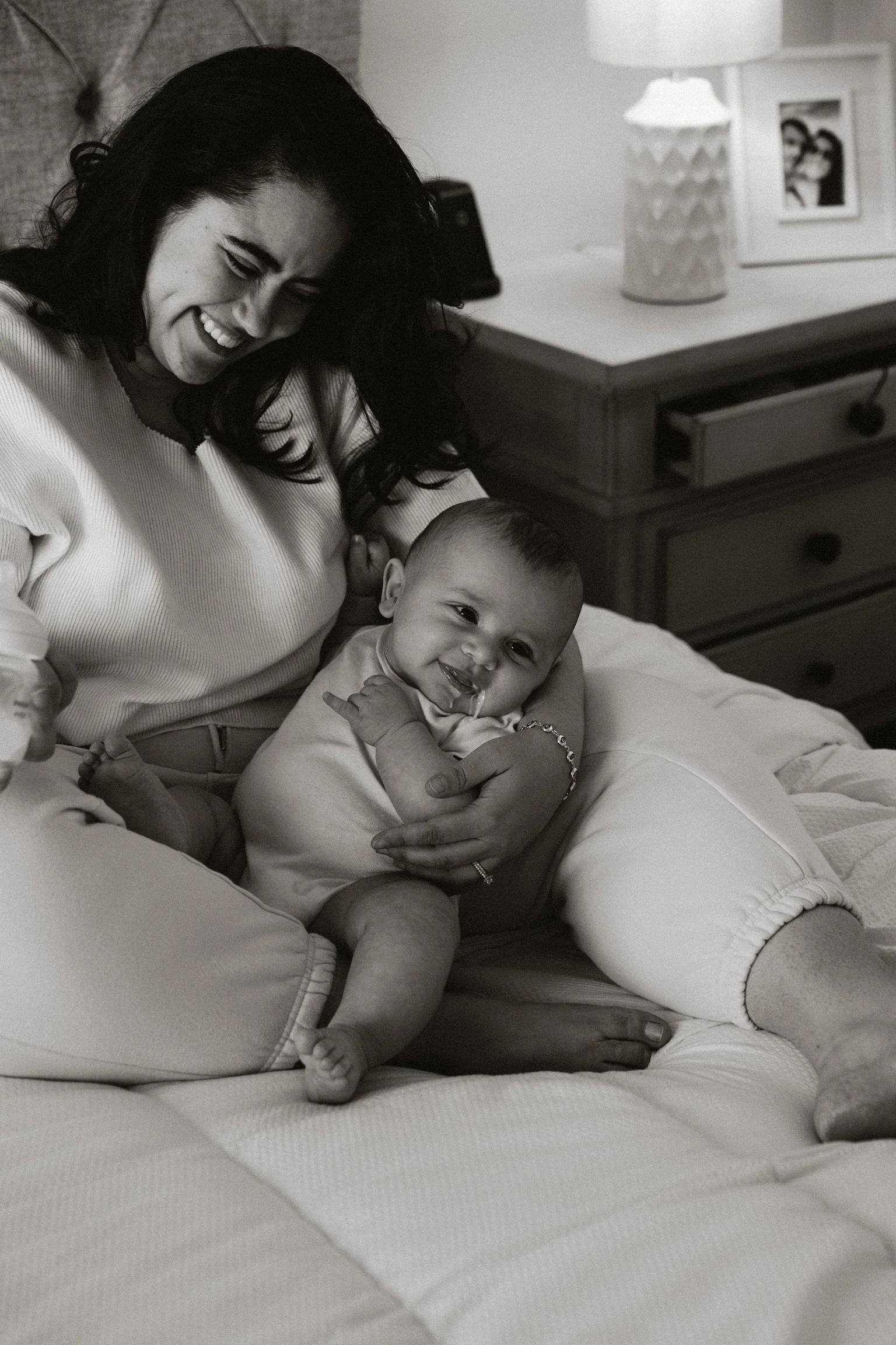 A woman holding a smiling baby on a bed, in a cozy room with a dresser, a lamp, and family photos in the background.