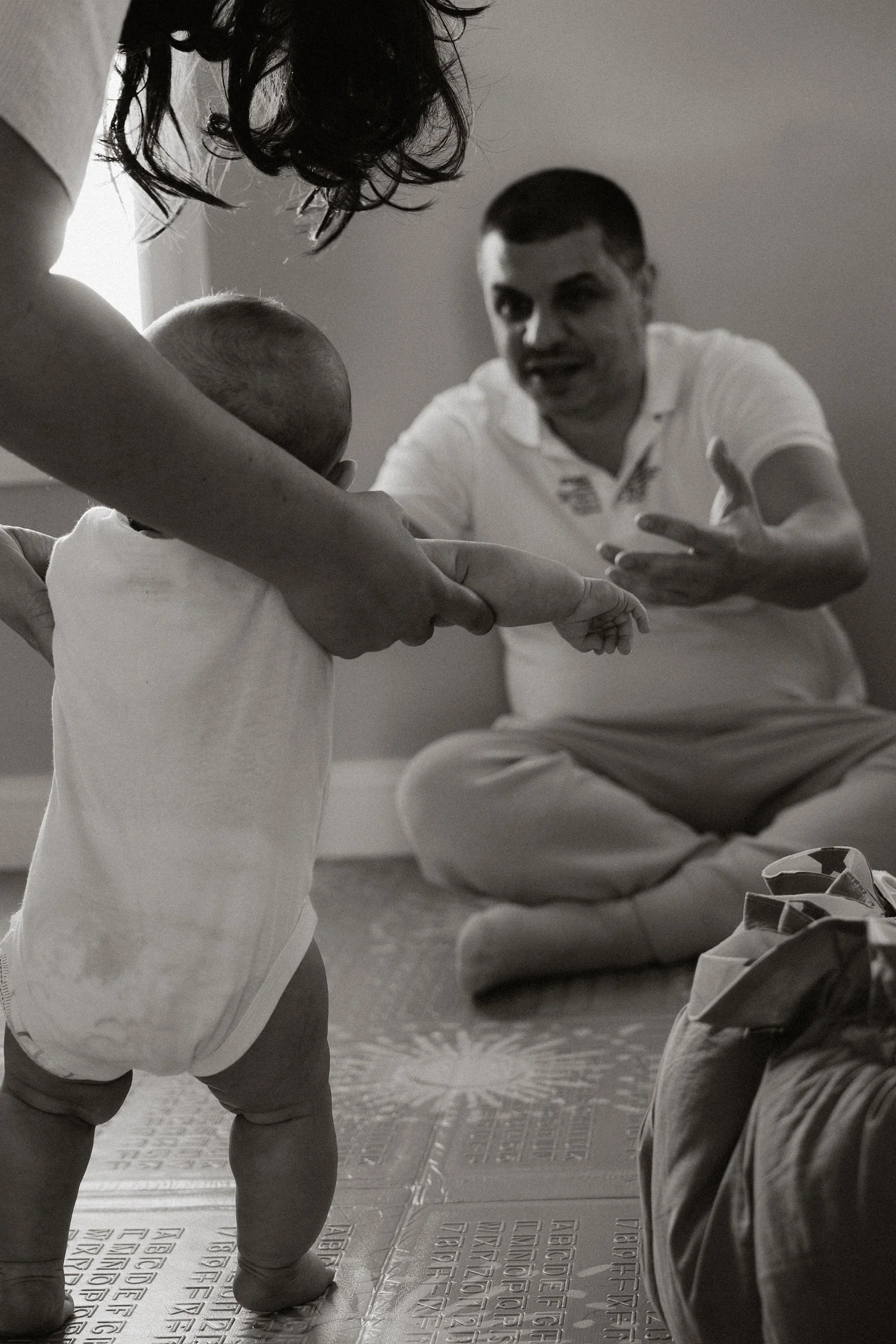 A man sitting on the floor with knees crossed, gesturing with his hands while talking to a woman and a baby. The woman is helping the baby stand, holding the baby's hands. The scene appears to be indoors with tanning sunlight coming through a window.