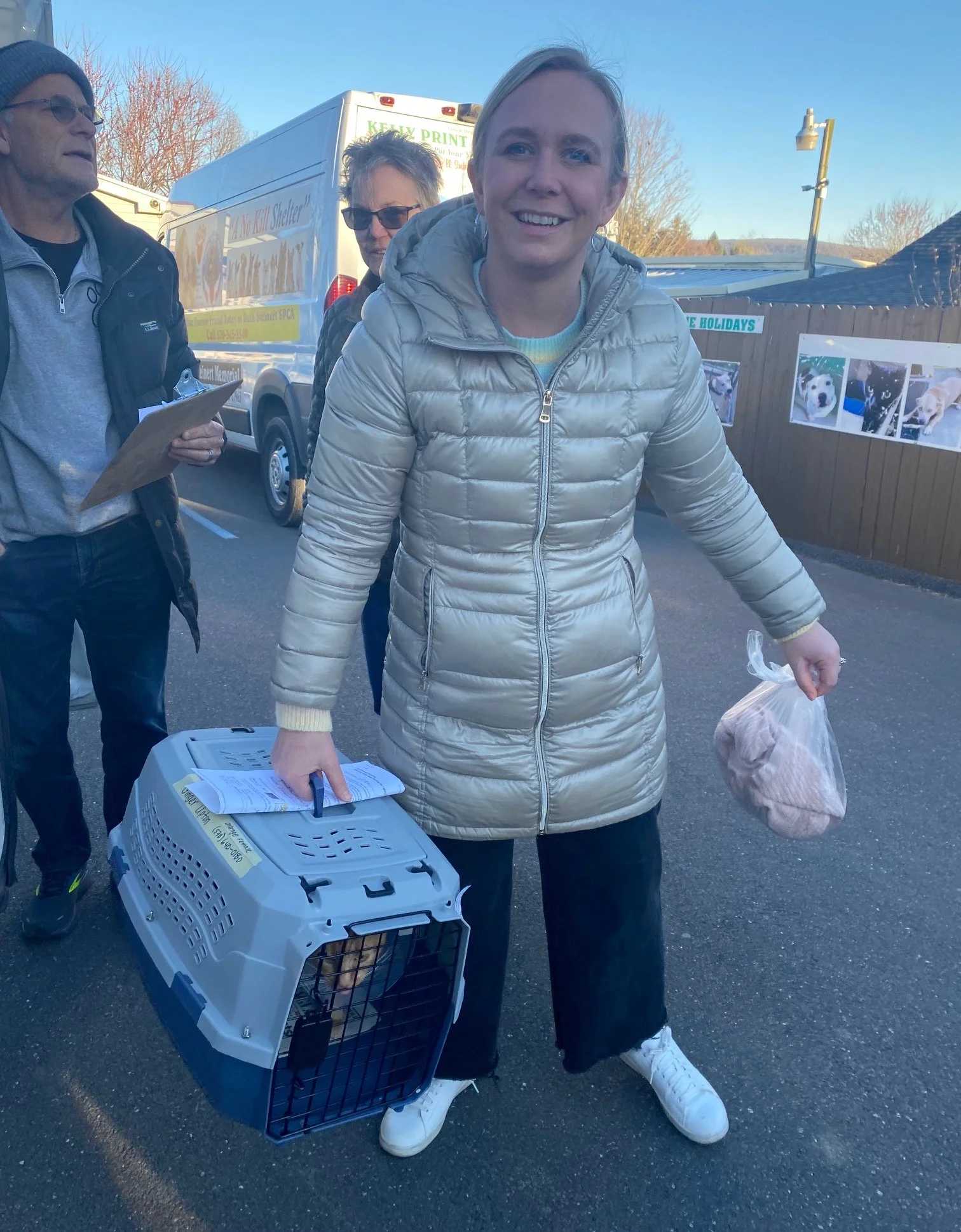 A smiling woman in a beige puffy jacket holding a plastic bag with a blanket or towel and a pet carrier with a small dog inside, standing outdoors in a parking lot.
