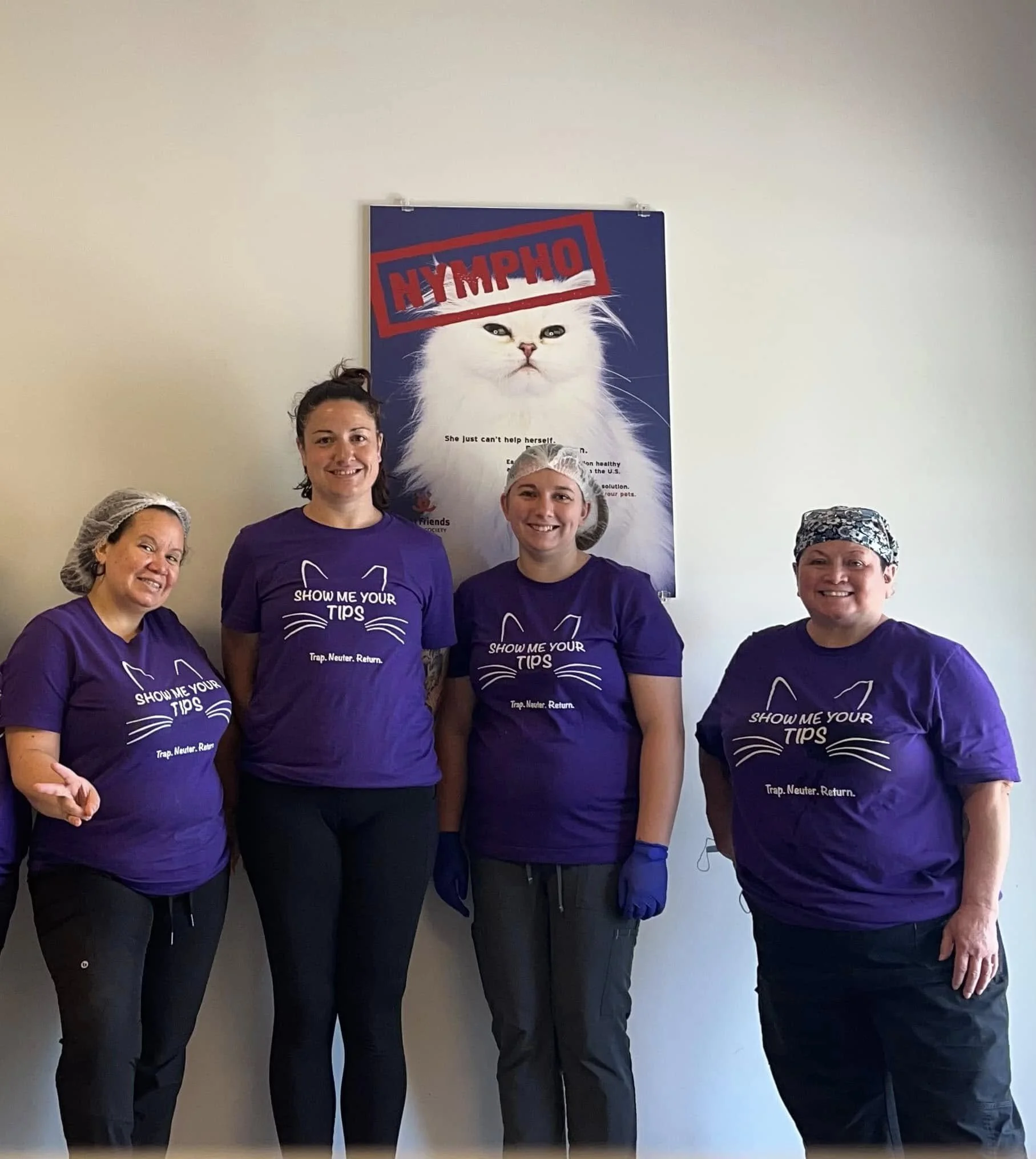 Four people in purple shirts and hairnets standing in front of a poster of a white cat with the word 'NYMPHO' printed over it.