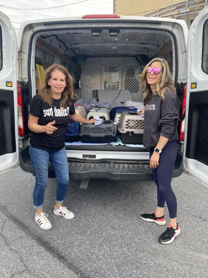 Two women smiling and standing next to the open back of a white van, which contains multiple pet carriers and supplies.