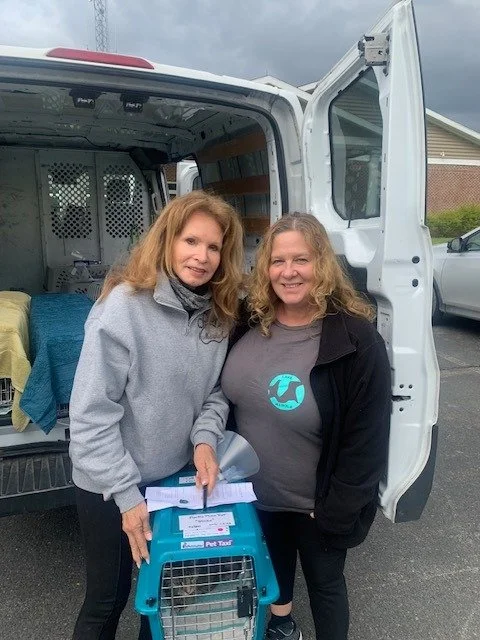 Two women standing outside next to a white van with its side door open. One woman is holding a pet carrier, and they are smiling at the camera.
