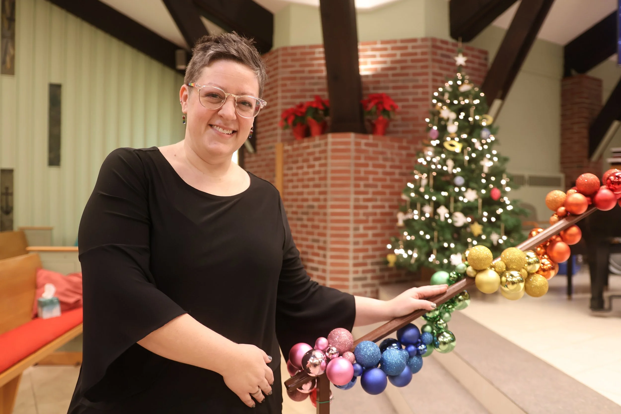 Person in black top smiling, resting hands on a hand rail wrapped with rainbow ornaments.
