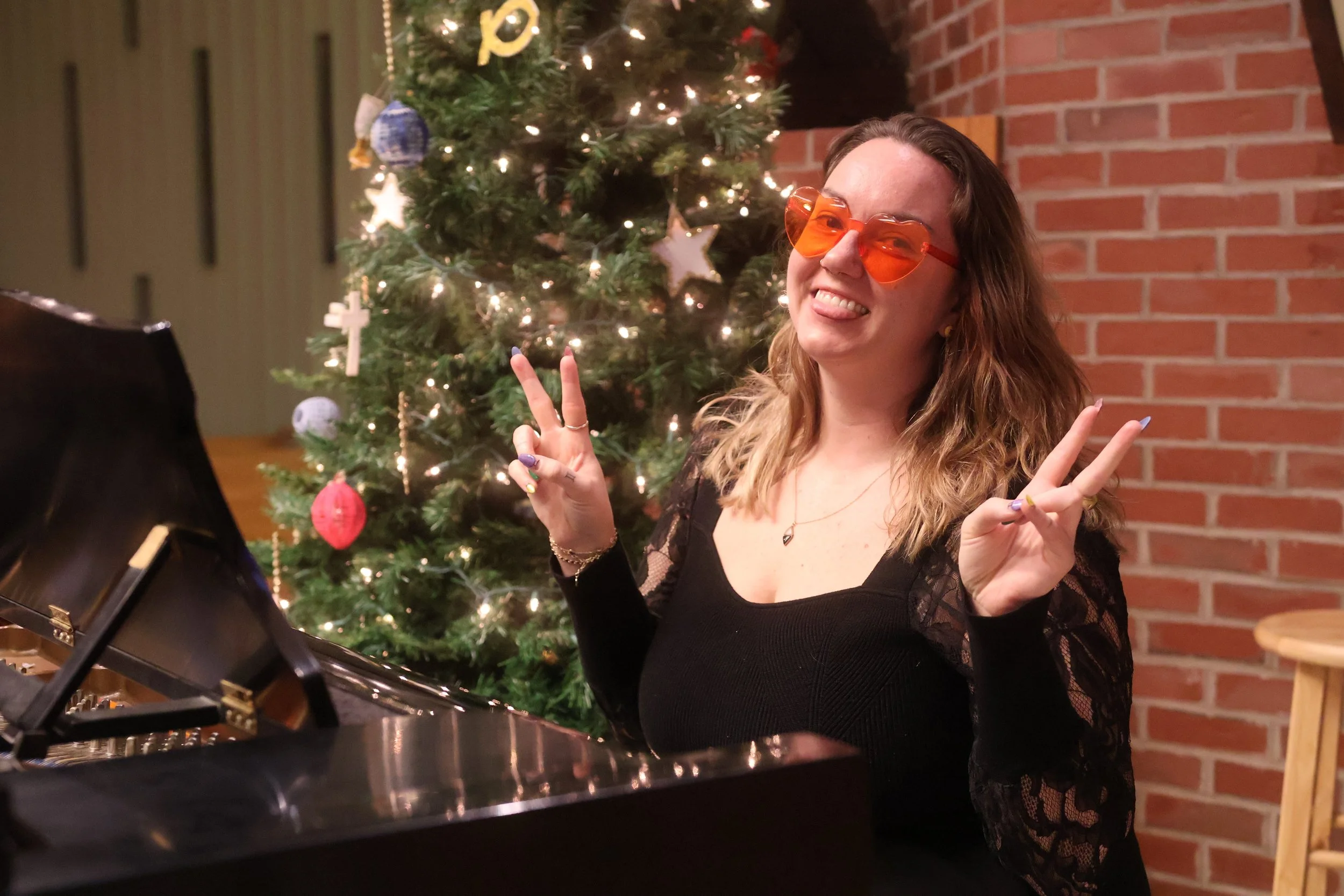 Woman sitting at black piano, smiling with her tongue out, wearing red heart sunglasses, making peace signs.