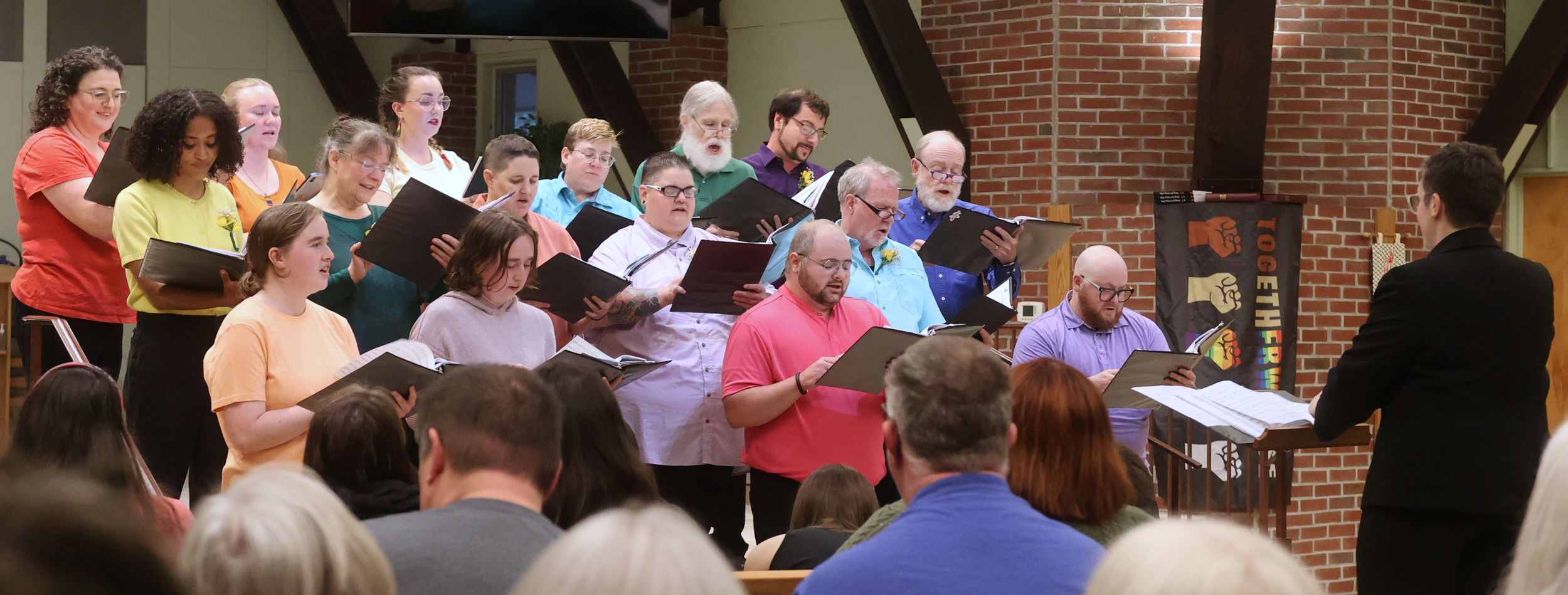 16 singers in colorful stops singing to an audience with a person in black conducting.