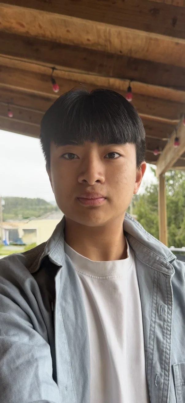 A young man with black hair wearing a gray jacket and white shirt taking a selfie outdoors under a wooden roof overhang, with trees, grass, and a building visible in the background.