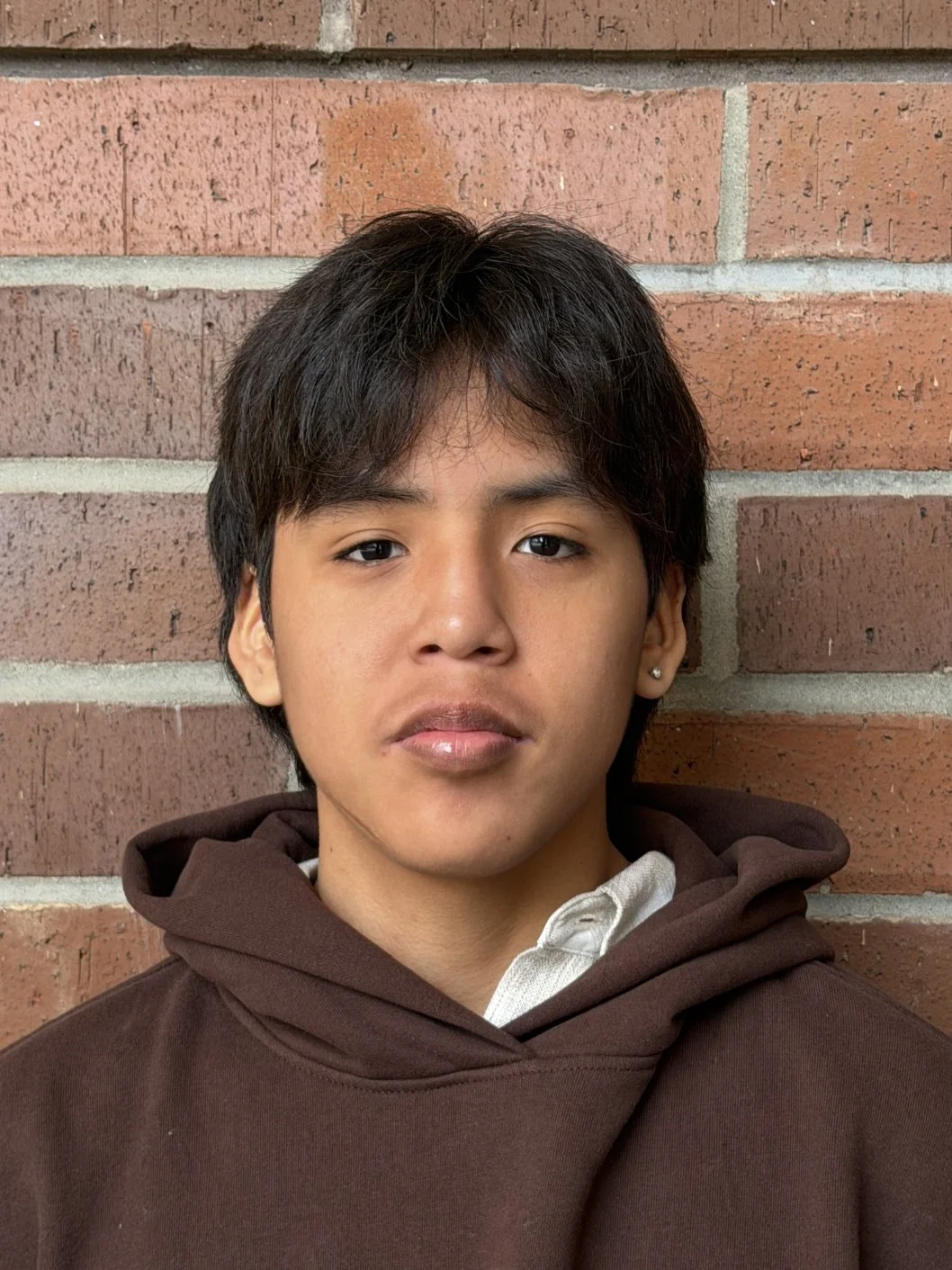 Close-up of a young person with black hair, wearing a dark brown hoodie, standing against a brick wall.