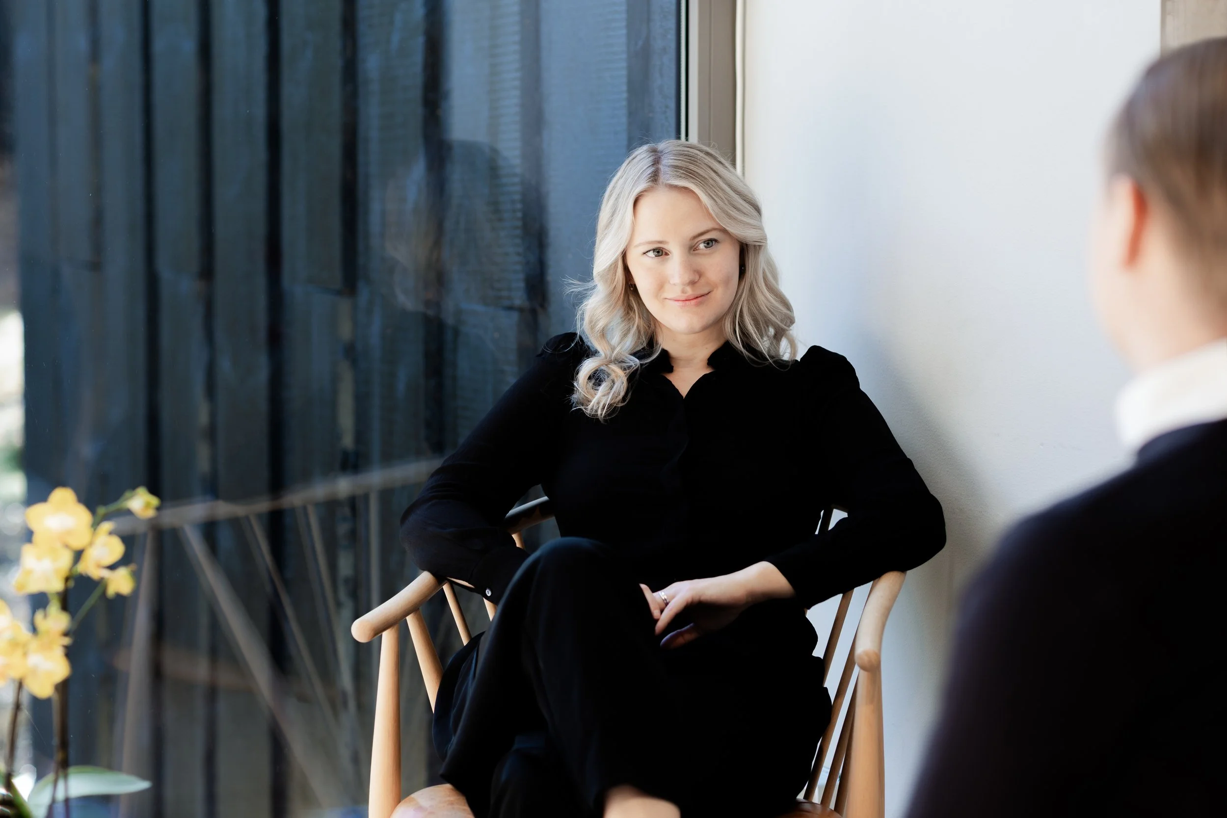 A woman with blonde wavy hair sitting on a wooden chair, engaged in conversation with a man whose back is to the camera, in a well-lit indoor space near a large window.