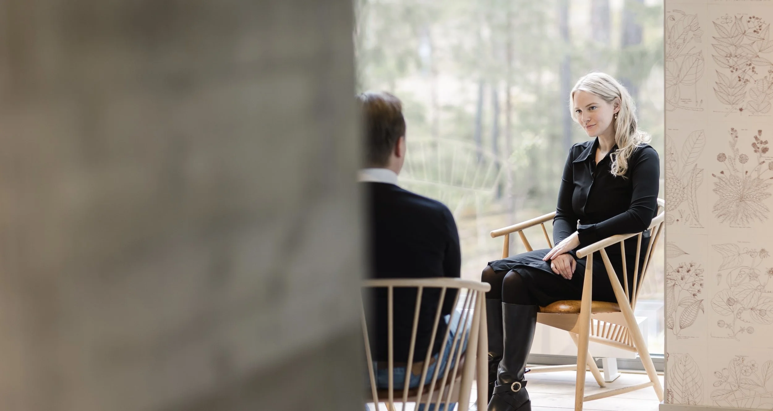 A woman with blonde hair sitting on a wooden chair, engaged in a conversation with a man whose back is visible, in a room with large windows and botanical wallpaper.
