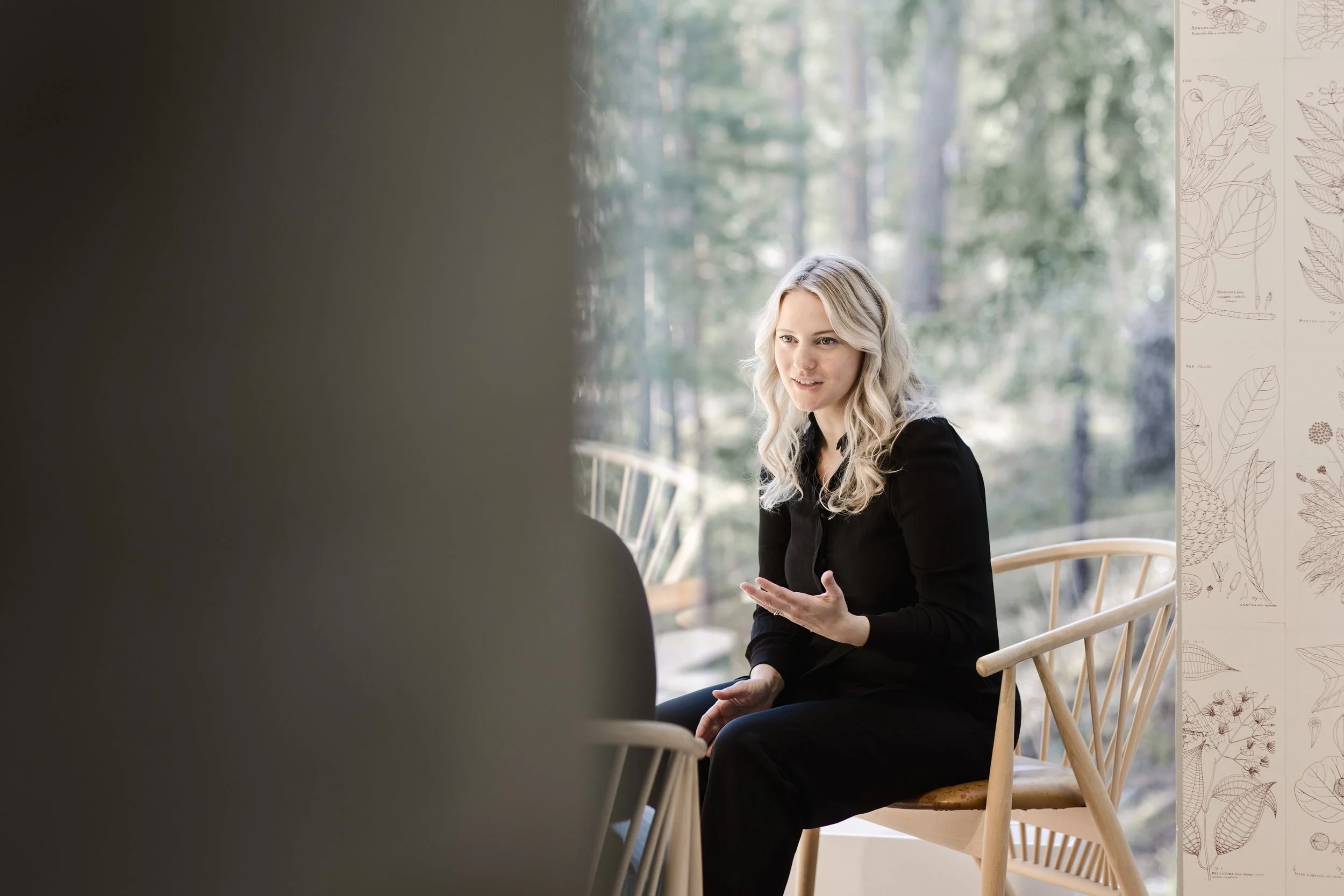 A woman with long blonde hair sitting on a wooden chair by a large window, engaging in conversation with someone whose back is visible in the foreground. The woman is gesturing with her right hand and appears to be explaining something. The background includes trees and a wall with botanical illustrations.