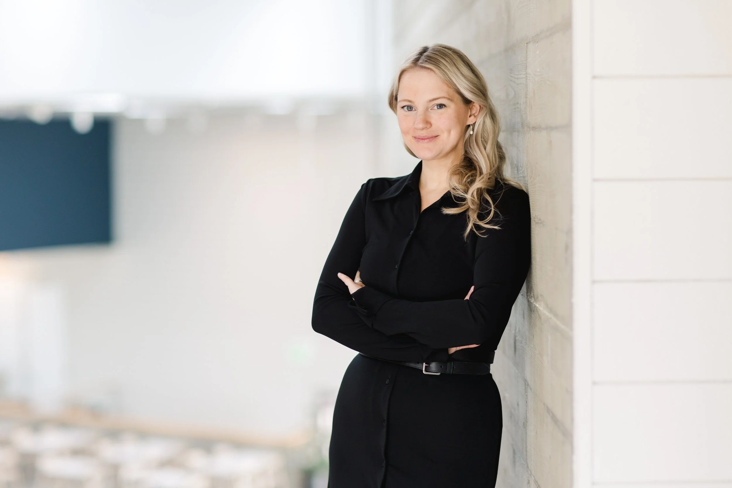 A woman with long curly blonde hair wearing a black shirt and black pants, standing with arms crossed, leaning against a light-colored wooden wall, smiling at the camera.
