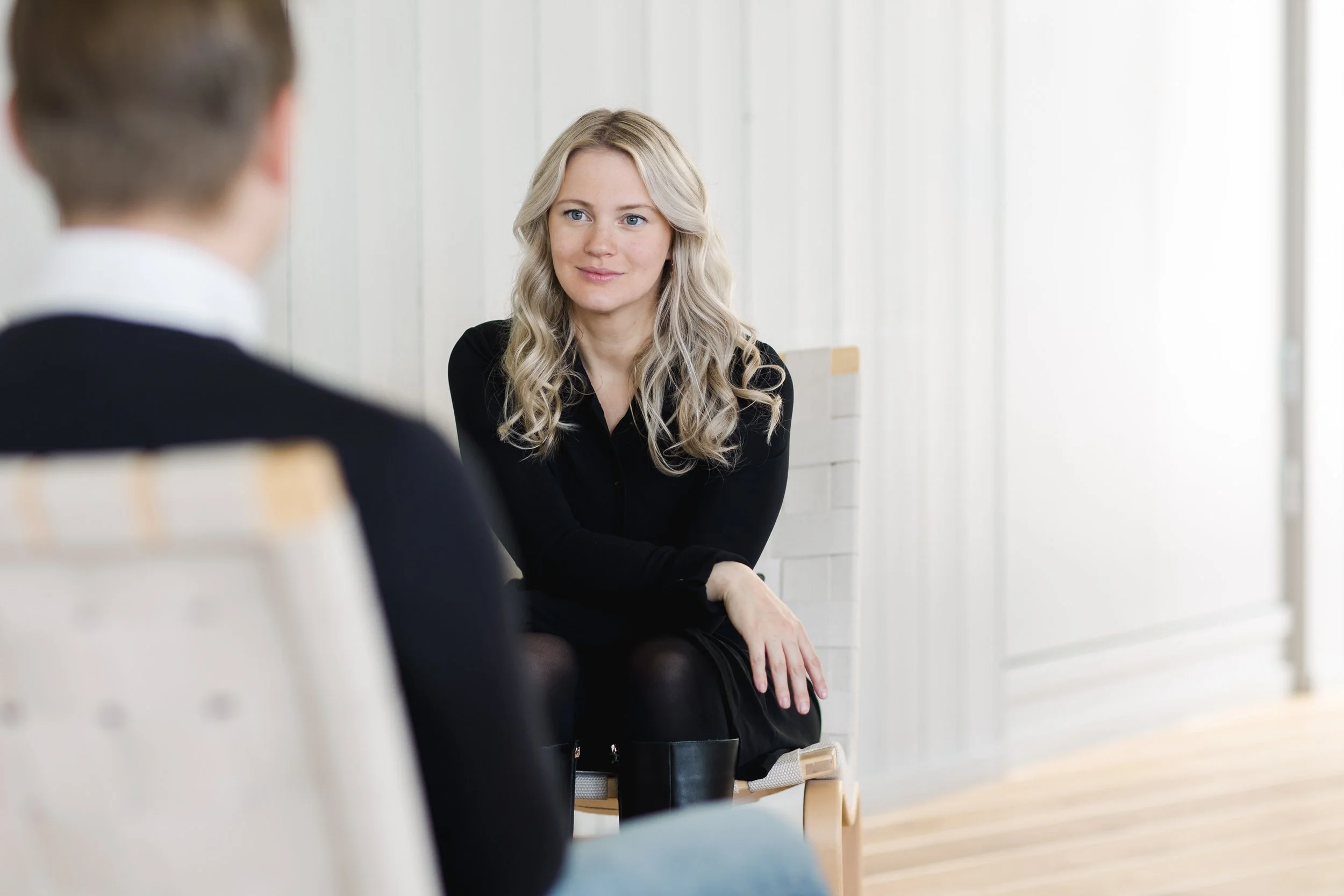A young woman with long blonde hair and blue eyes sitting and listening during an interview or conversation with a man whose back is to the camera, in a bright room with white walls.