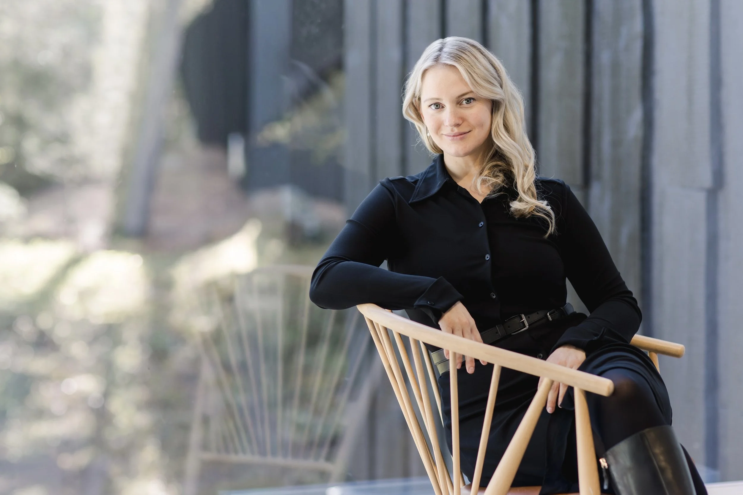 A woman with blond, wavy hair and blue eyes sitting outdoors, wearing a black button-up shirt and black belt, leaning on a light wooden chair with a modern design.