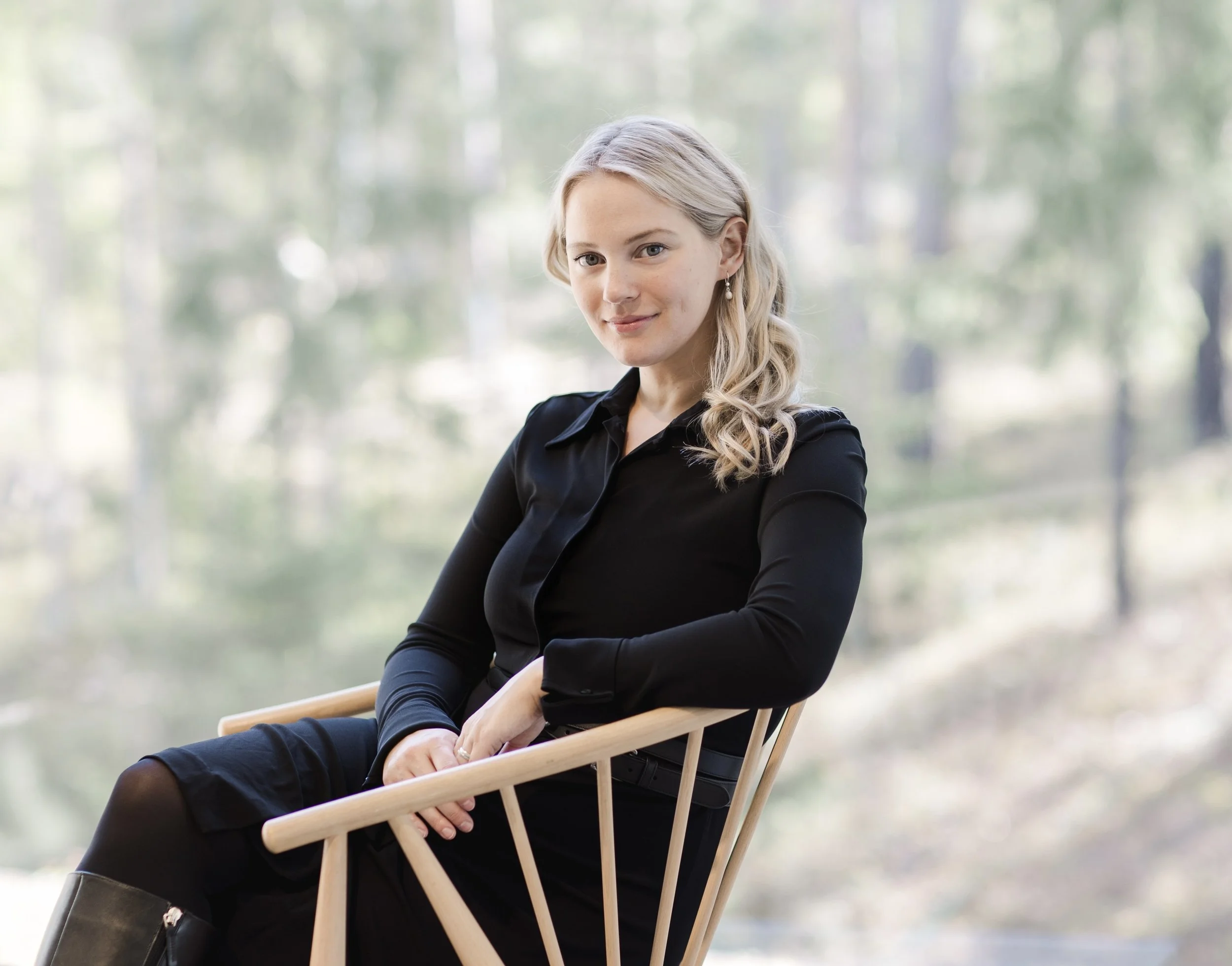A young woman with blonde hair, wearing a black dress and jewelry, sitting outdoors on a wooden chair with a scenic background of trees and soft sunlight.