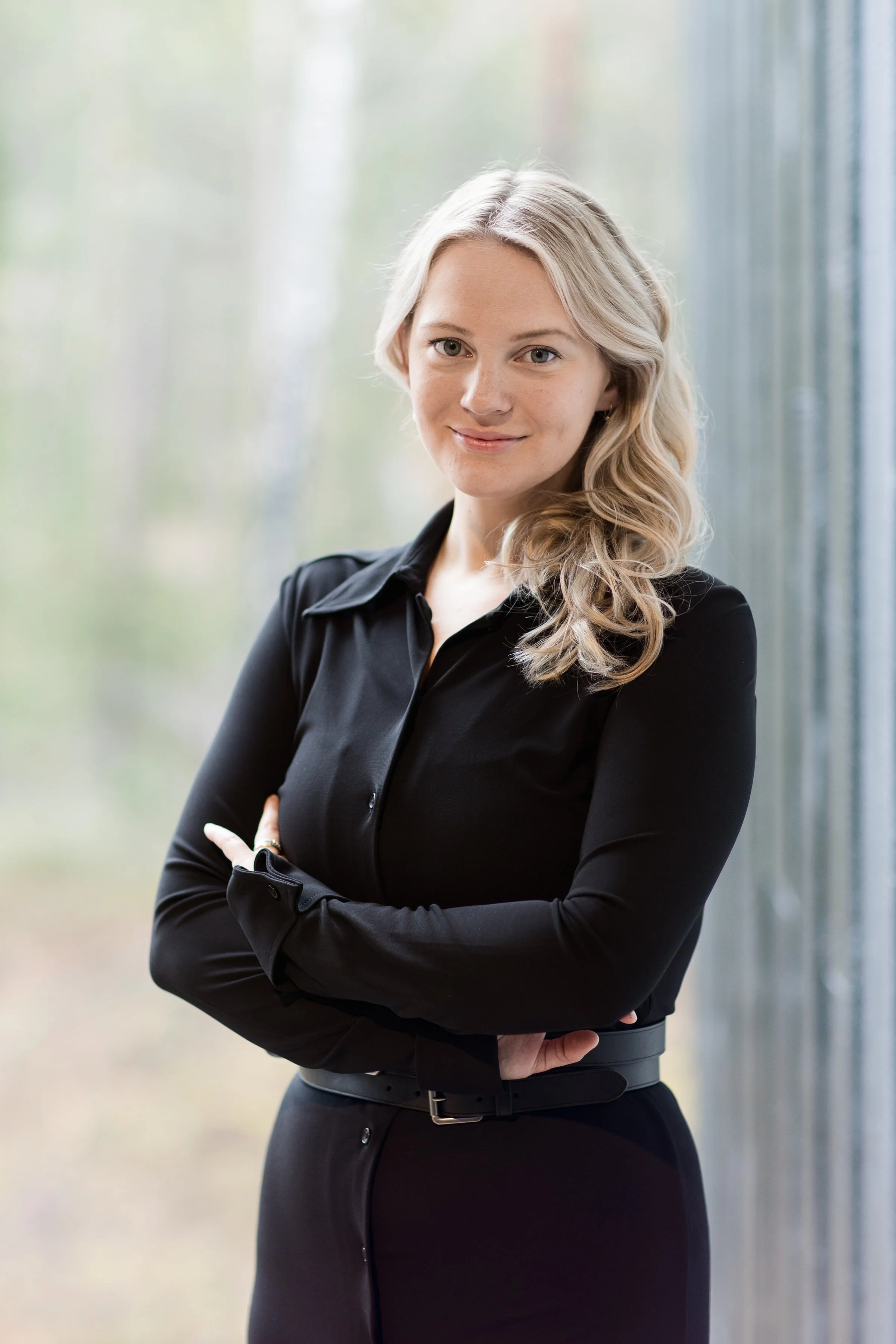 A woman with blonde, wavy hair and fair skin, smiling, wearing a black long-sleeve blouse, with arms crossed, standing in front of a blurred background.