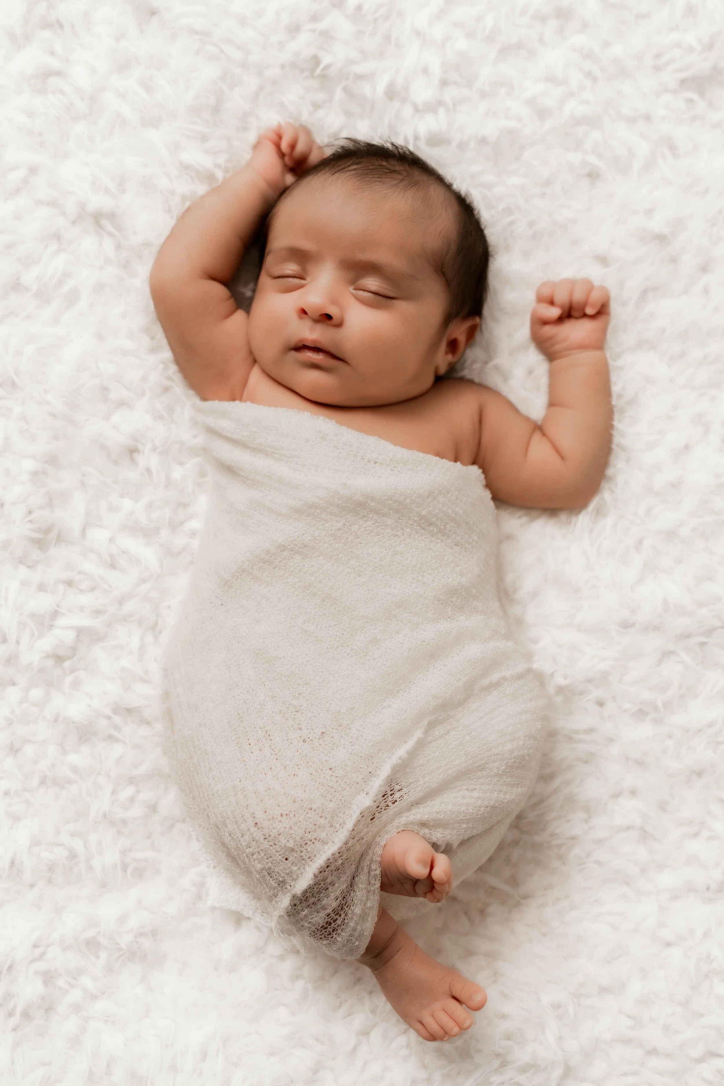 A newborn baby sleeping on a fluffy white blanket, wrapped in a light beige knitted blanket, with arms raised and eyes closed.