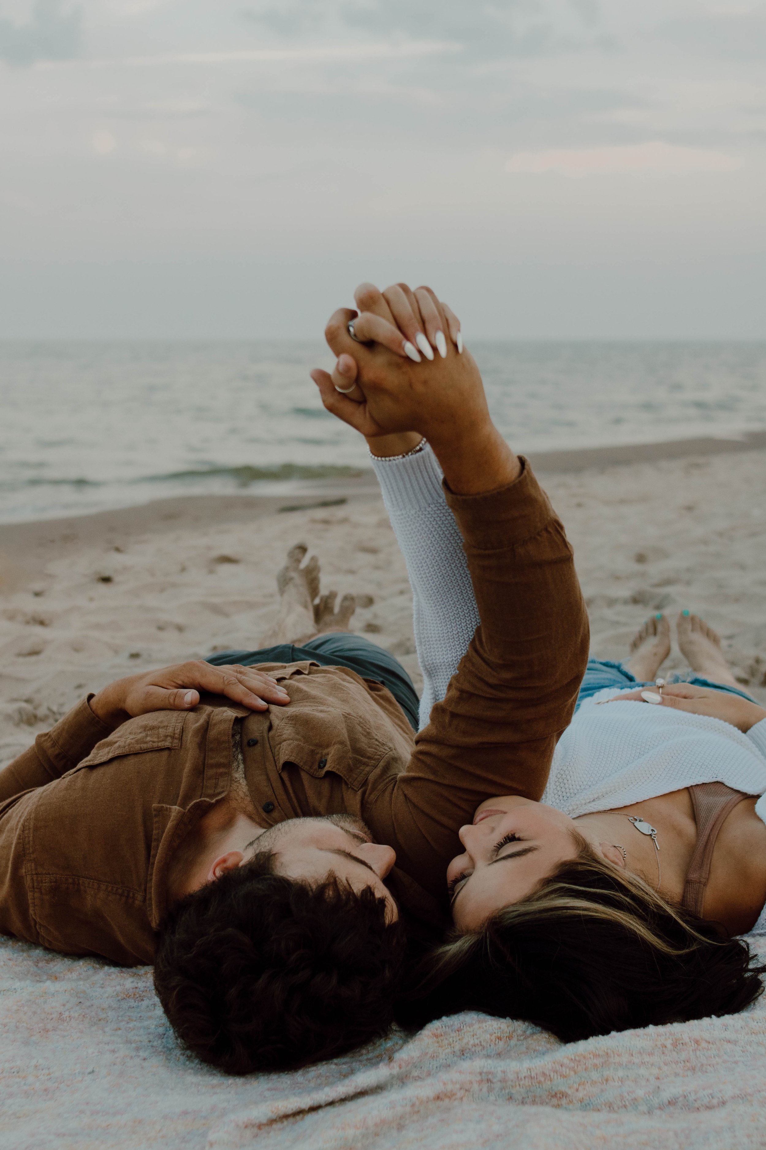 A couple lying on the beach, holding hands intertwined, with the ocean and cloudy sky in the background.