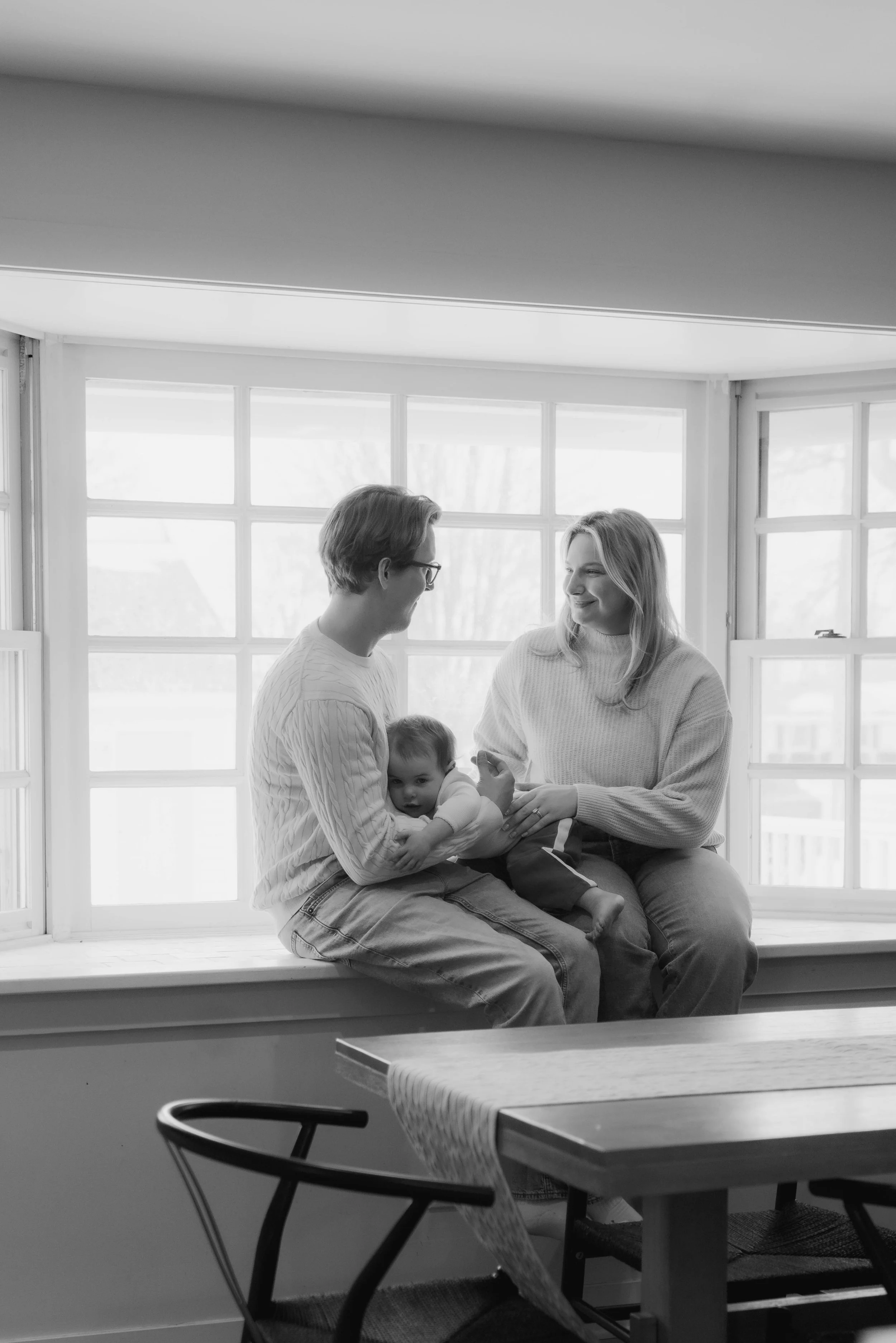 A family of three sitting by a large window, with a man, a woman, and a baby, enjoying a happy moment together.