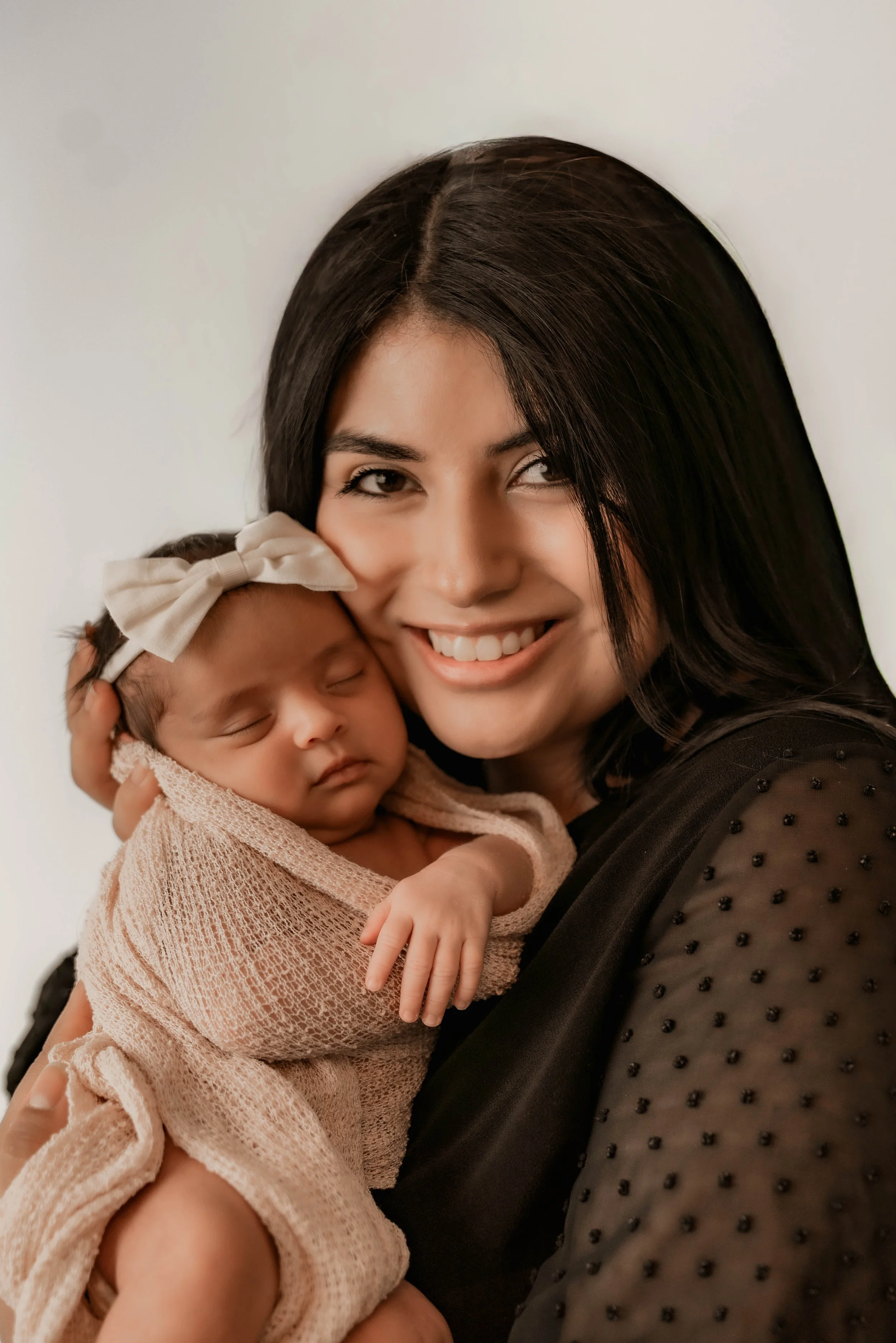 A woman with long black hair holding a sleeping baby girl wrapped in a pink blanket and wearing a white bow headband, smiling at the camera.