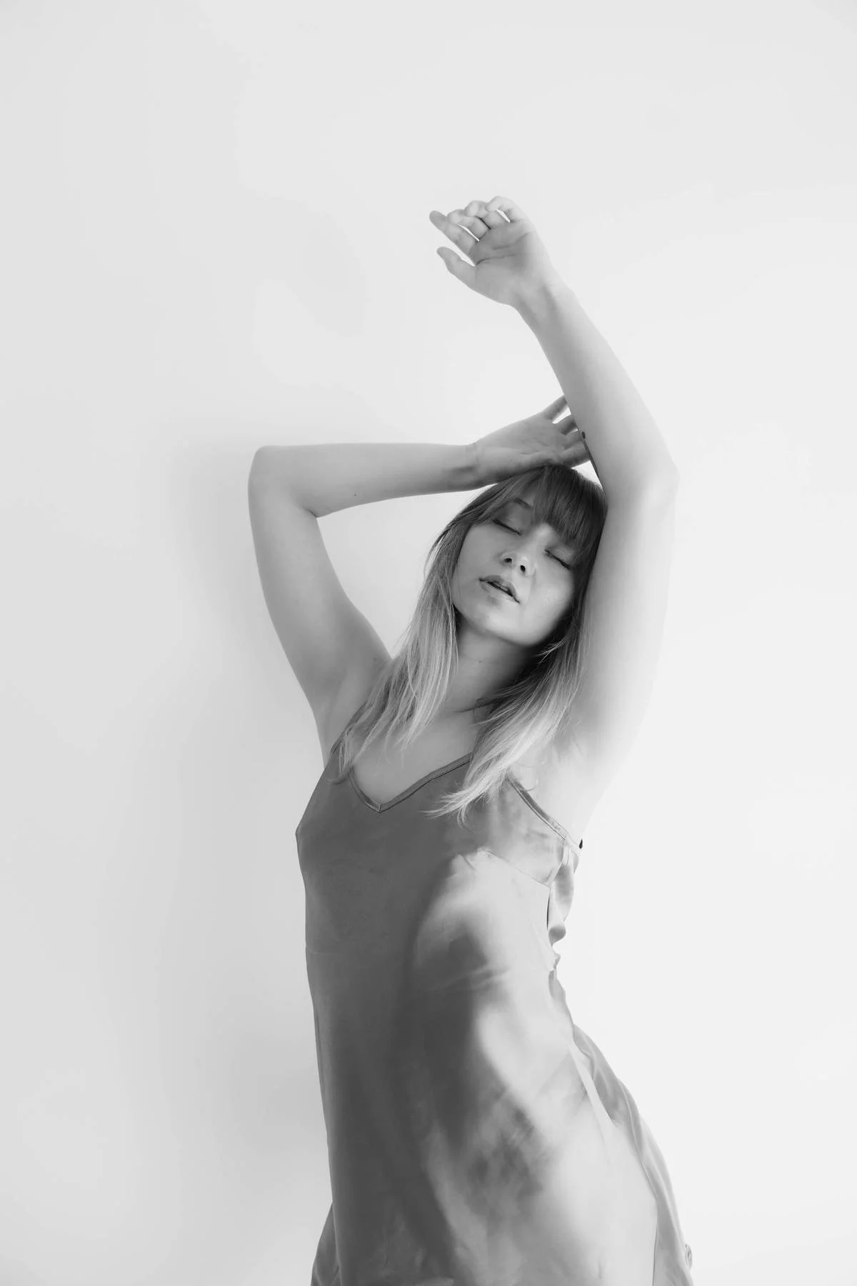 A woman in a silky dress with closed eyes, one arm raised above her head, and the other arm bent, touching her head, against a plain background.