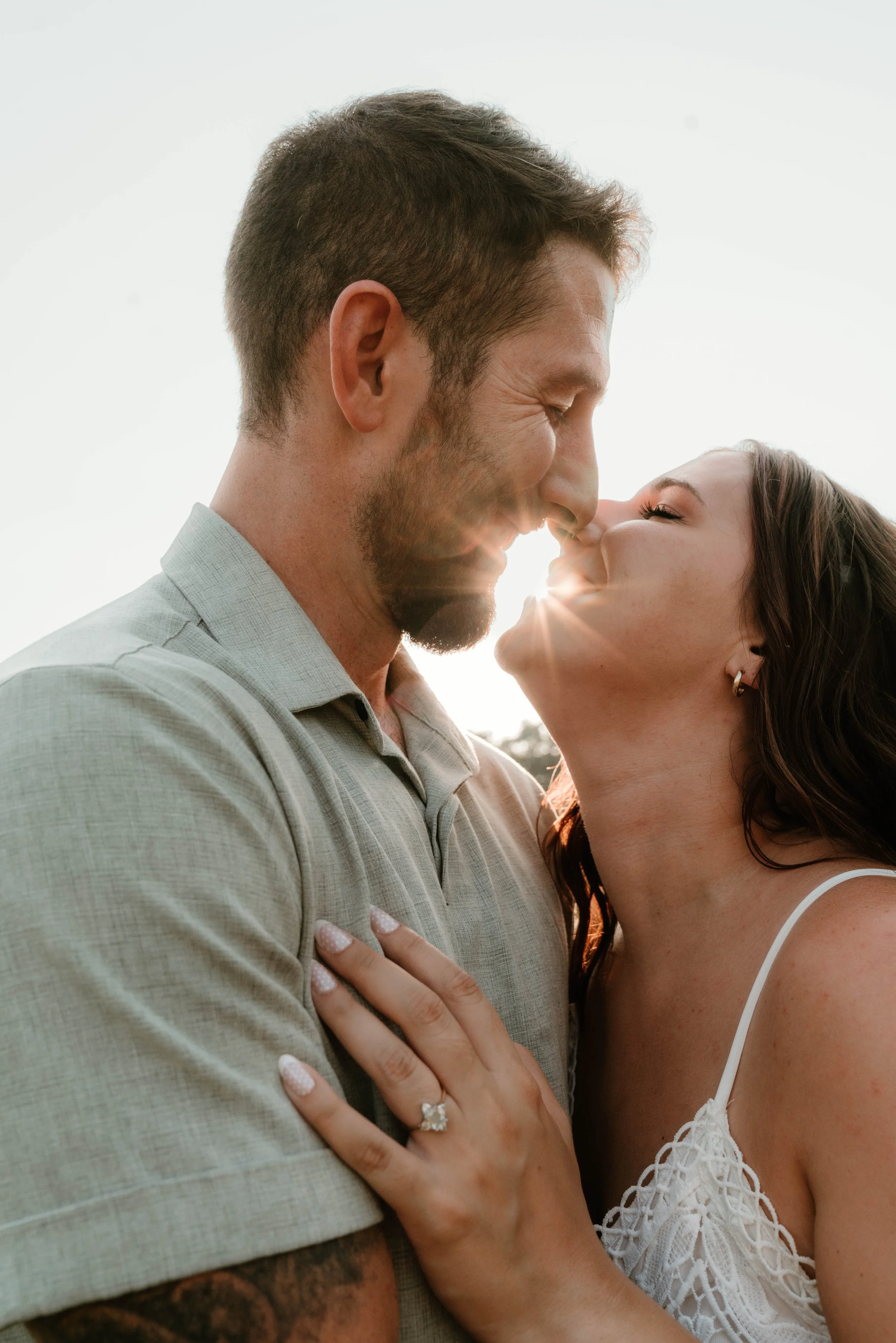 A couple is about to kiss outdoors during sunset, with the sun visible between their faces.